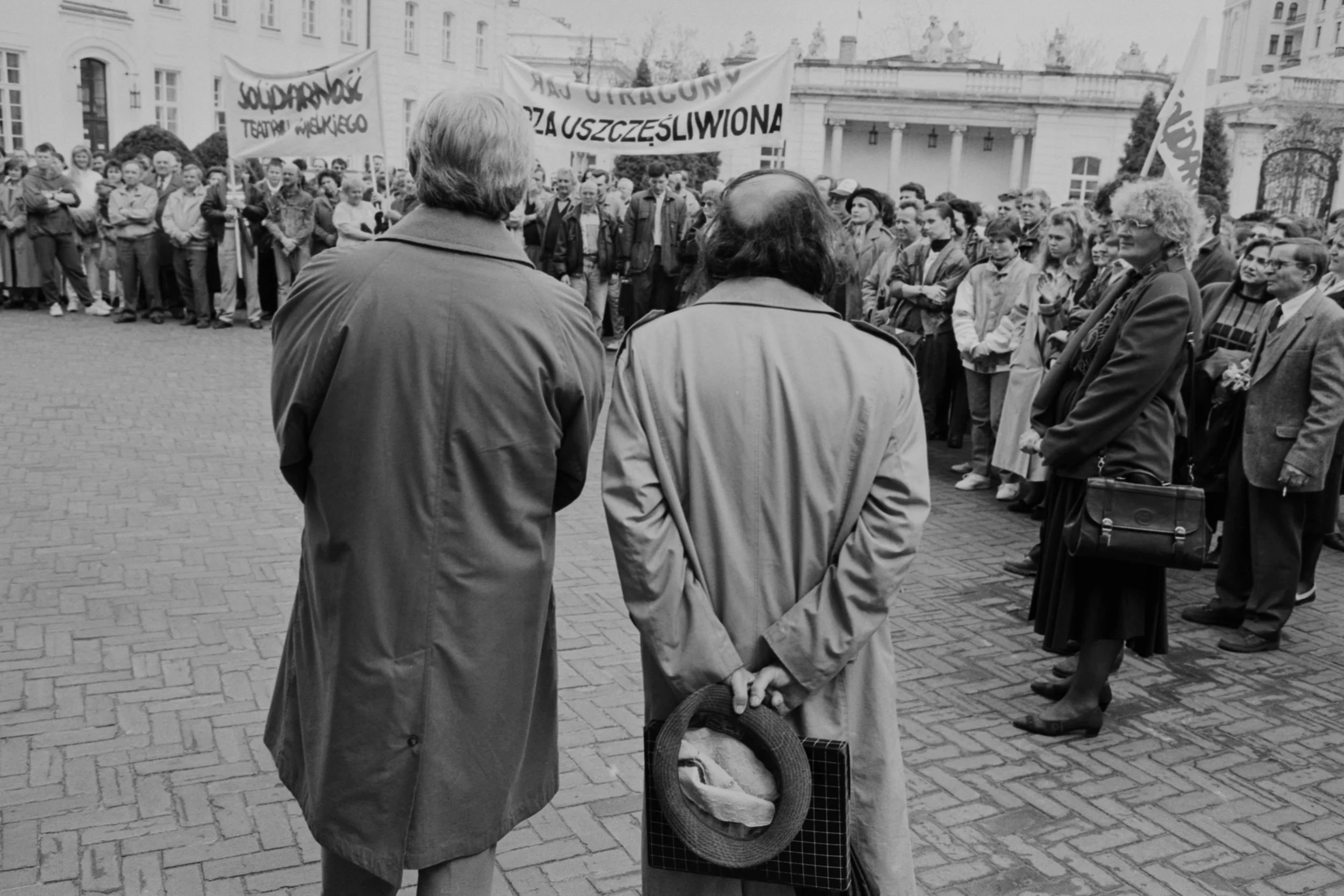 A black and white photo of a crowd gathered outdoors, with two women in the foreground seen from behind. One woman holds a hat and a small bag, while the crowd holds banners and signs, indicating a demonstration or rally.