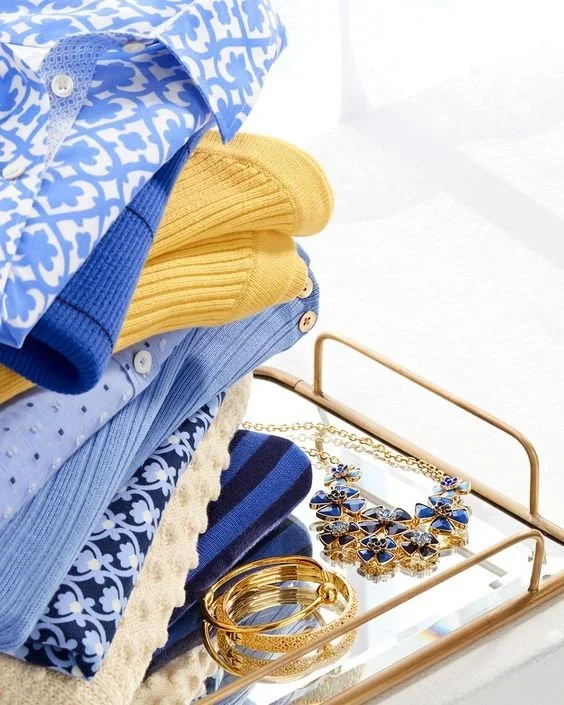 Stacked folded clothes in yellow and blue with a jewelry set of gold bracelets and a blue and gold necklace on a glass and gold tray.