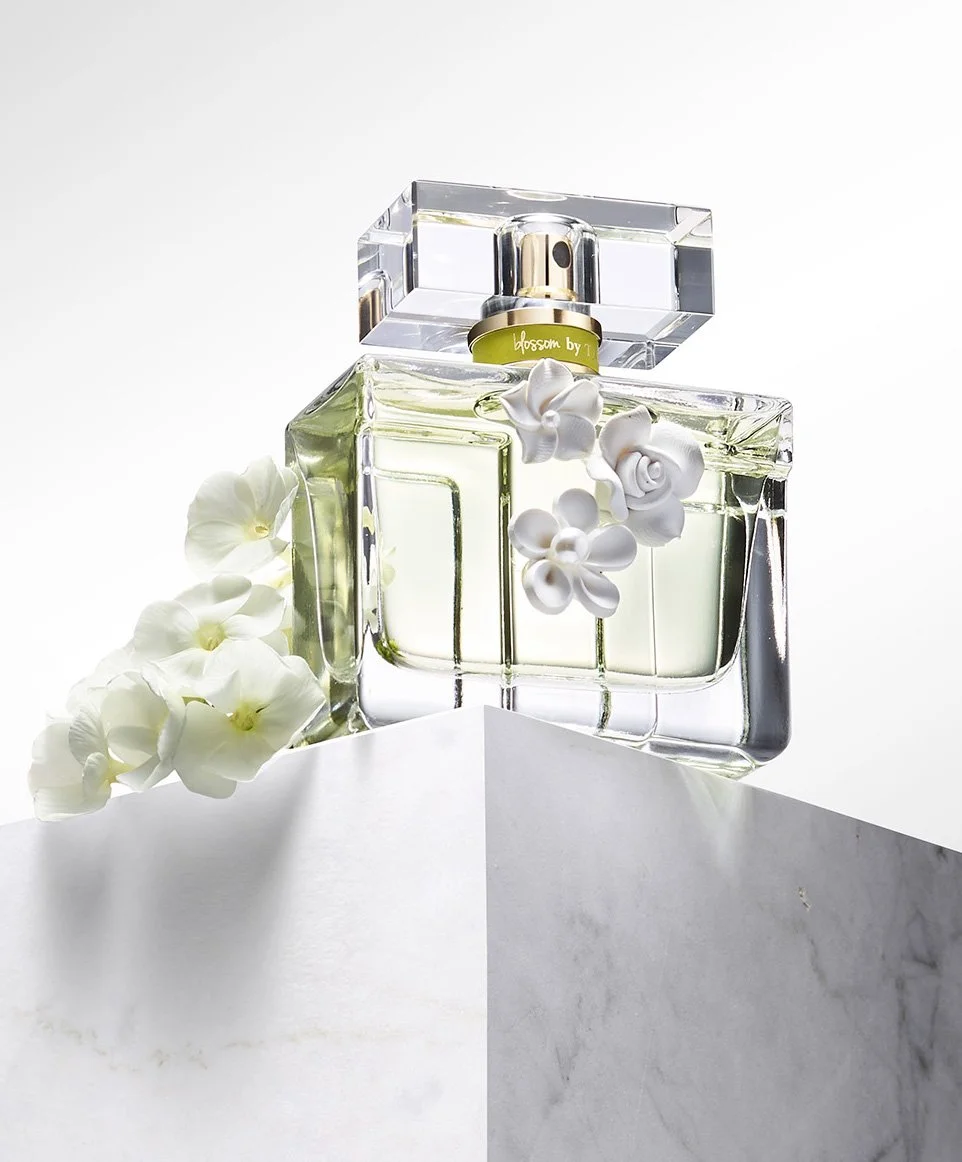 A perfume bottle with light green liquid, decorated with white flowers, placed on a white marble surface with a plain light background.