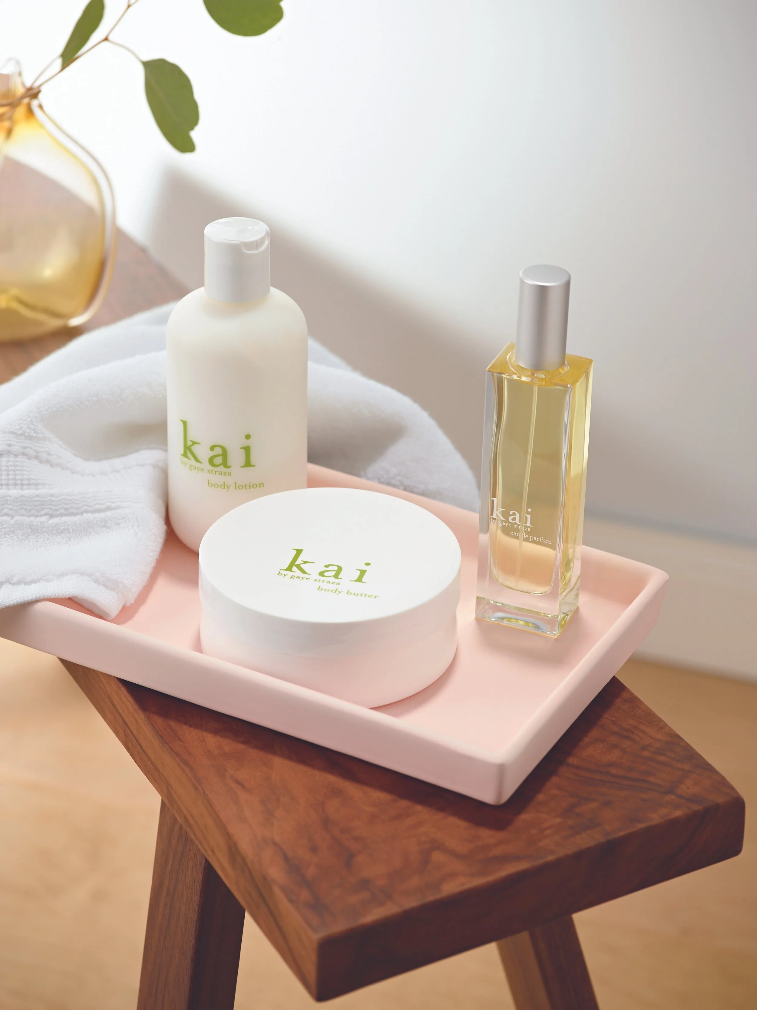 A pink rectangular tray on a wooden table holding skincare and fragrance products, including a white bottle labeled kai body lotion, a round white container labeled kai body butter, and a tall rectangular clear perfume bottle labeled kai eau de parfu