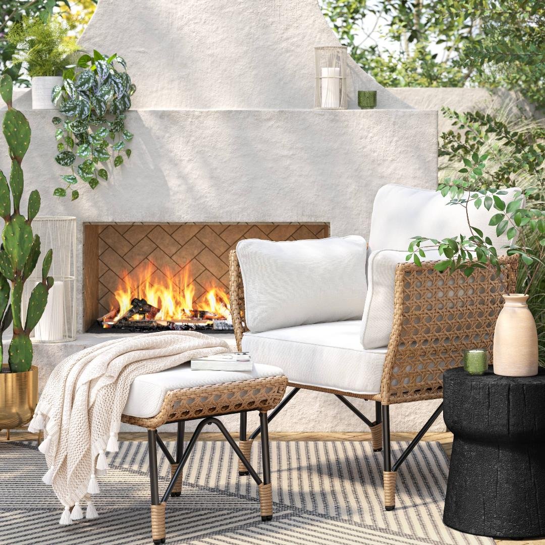 Cozy outdoor seating area with a white cushioned chair, a matching ottoman with a blanket, plants including a cactus, and a fireplace with a fire burning, surrounded by green foliage and decorated with candles and pottery.