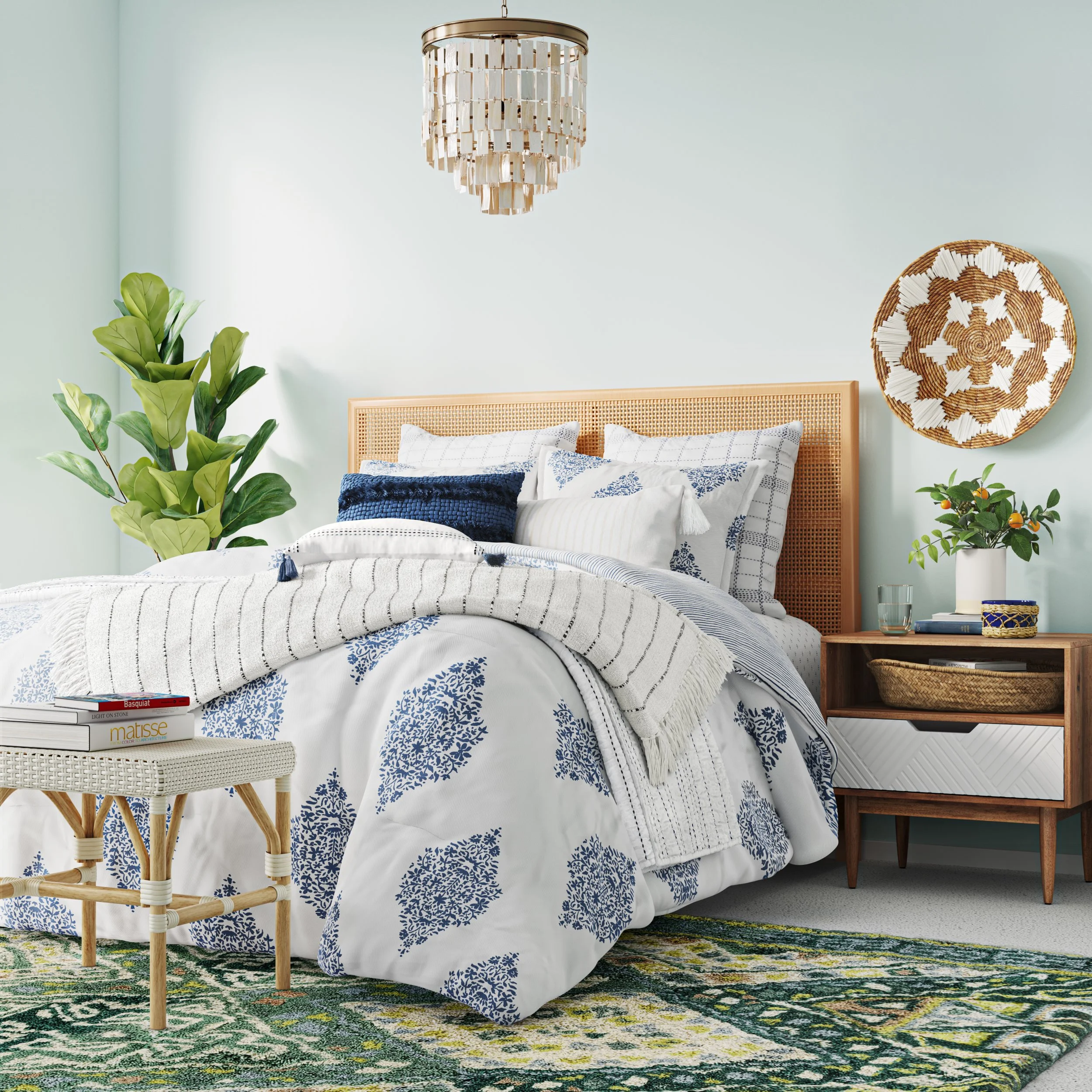 A neatly made bed with white and blue patterned bedding, multiple pillows, and a light wooden headboard in a bright room. To the right, a nightstand with a potted plant, glass of water, and decorative basket. On the wall, a woven round wall hanging. 