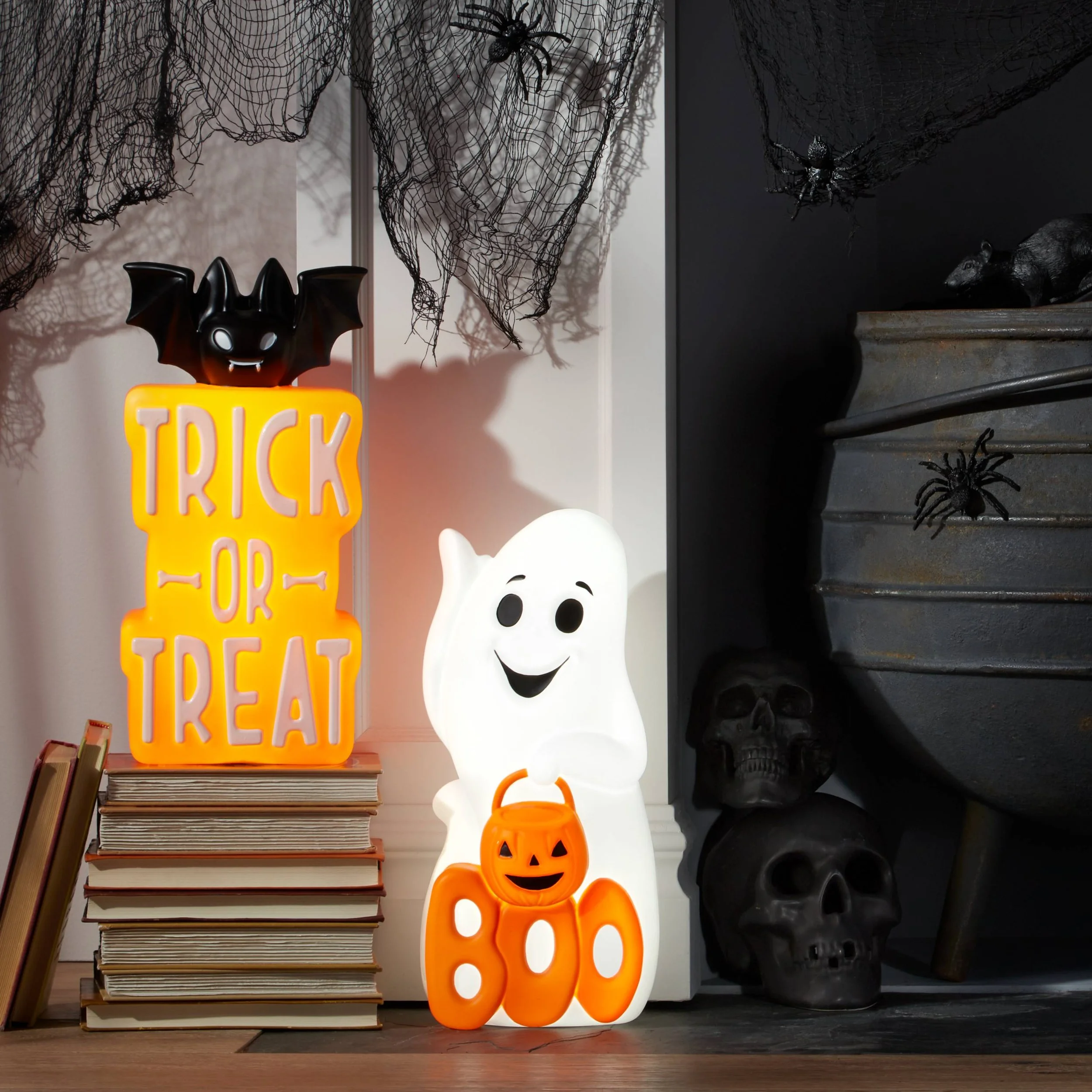 Halloween decorations including a lit sign that says 'Trick or Treat' with a bat on top, a smiling ghost figure, a small pumpkin container, stacked books, black skulls, spider webs, and spider decorations.