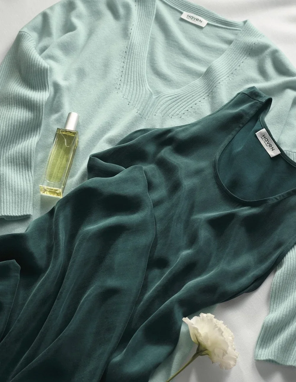 Light blue and dark green clothing laid out with a bottle of perfume and a white flower.
