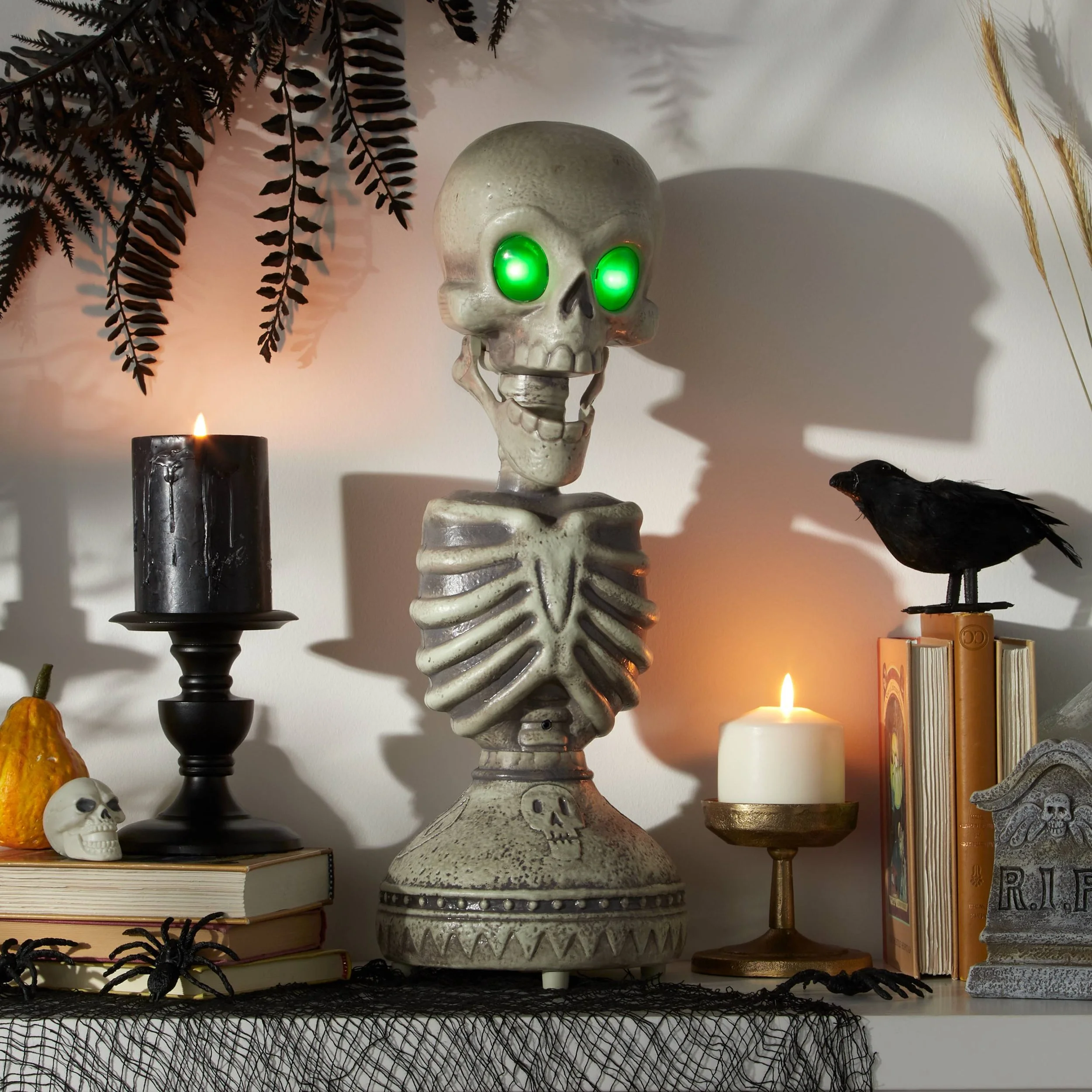 Decorative Halloween display with a skeleton skull with glowing green eyes, a black candle on a candlestick, a small skull, a yellow gourd, and a black spider on books, with candles casting shadows, autumn leaves, and a raven figurine on a pile of bo