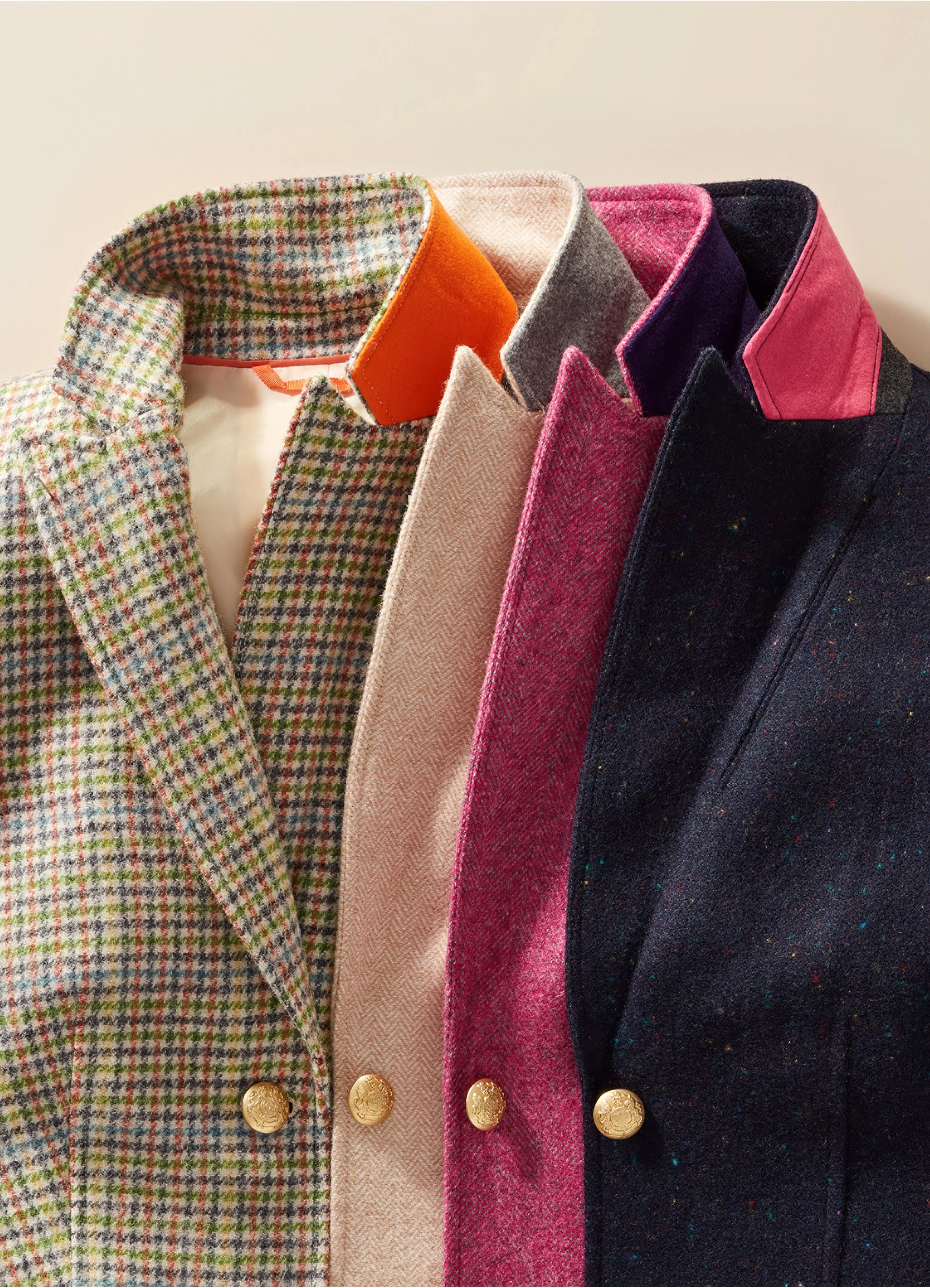 Four blazers hanging side by side, each with gold buttons. The first blazer has a colorful checkered pattern, the second is beige with a textured pattern, the third is pink with a herringbone pattern, and the fourth is dark navy with colorful specks.