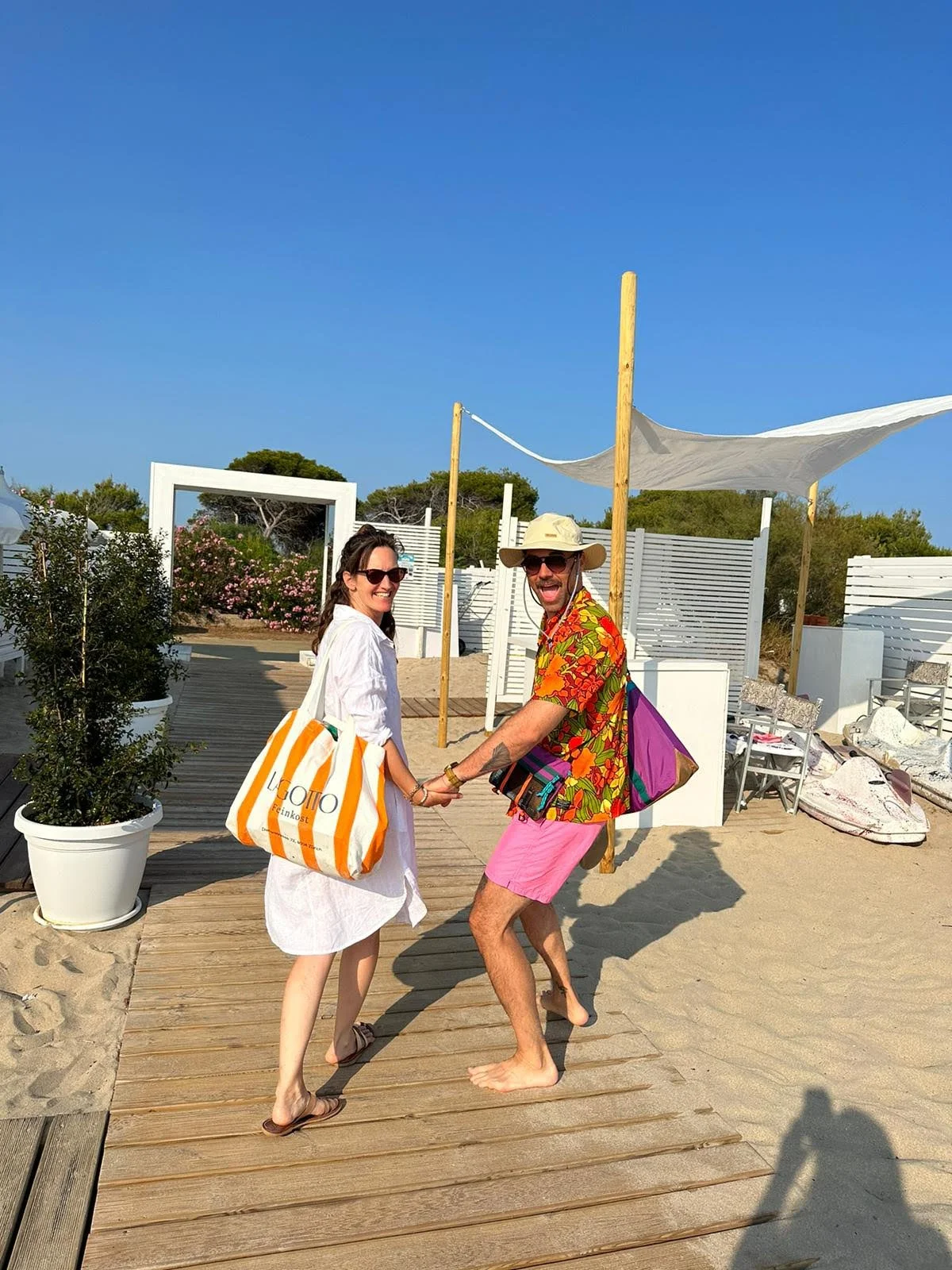 Ein Paar, das am Strand Spaß hat, hält Händchen und lacht, im Hintergrund Strandliegen und strahlend blauen Himmel.