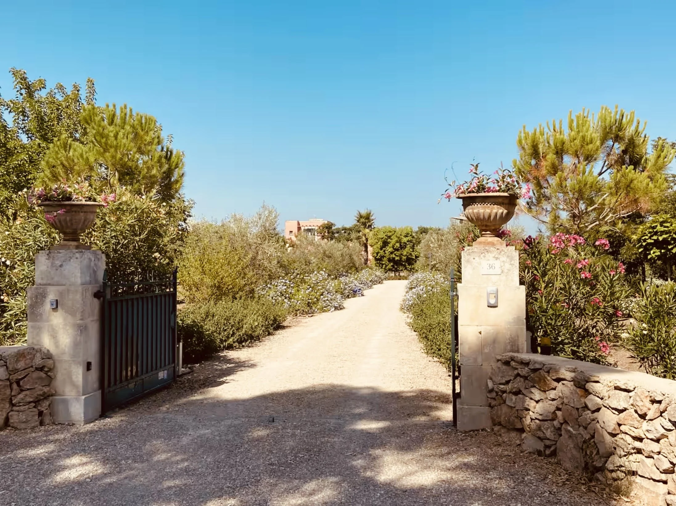 Ein Gartenweg, flankiert von Steinmauern und blühenden Pflanzen, mit eines Wachtürmen und einem blauen Himmel im Hintergrund.