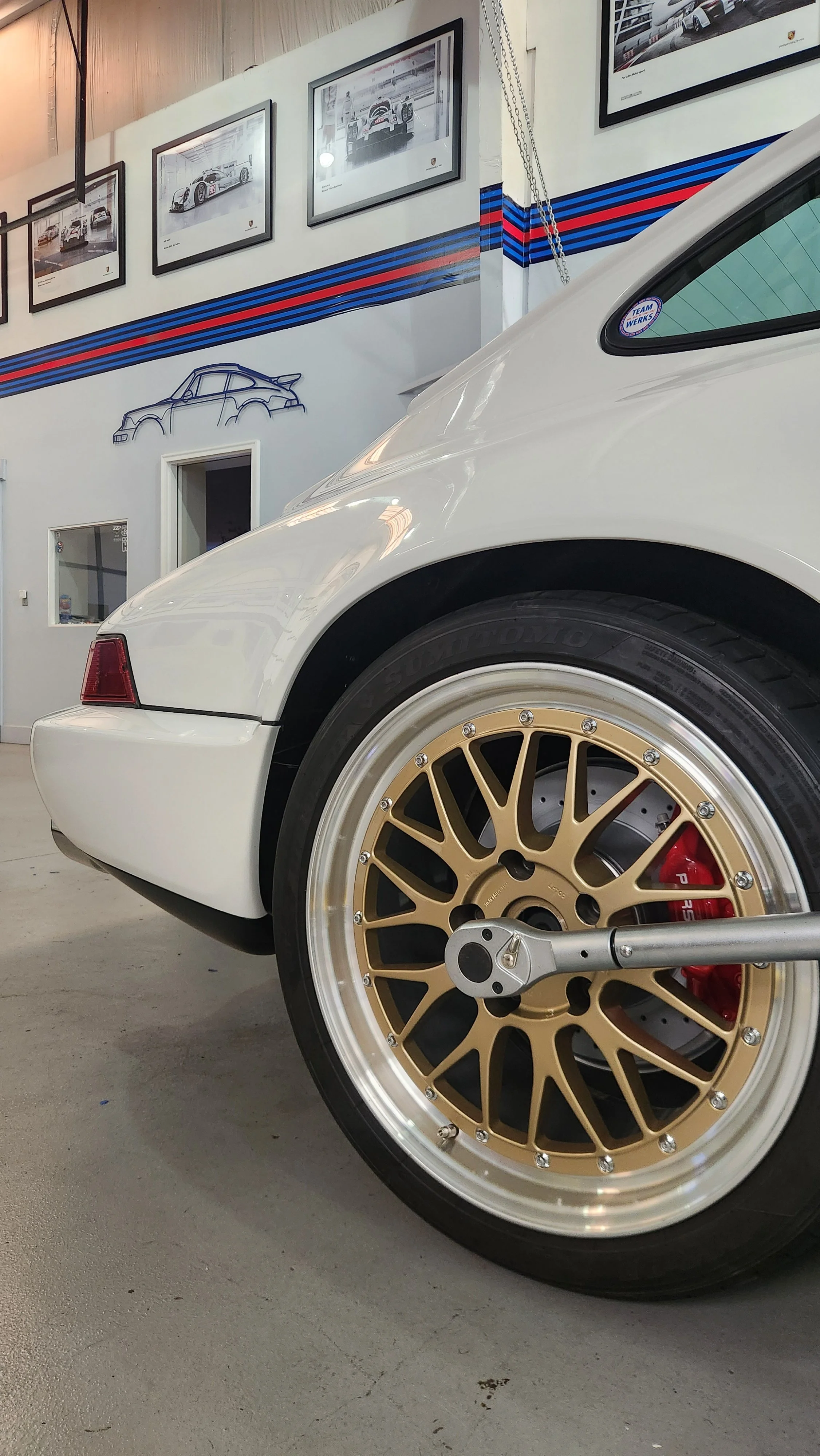 Close-up of a white Porsche 911 with gold BBS wheels and red brake calipers and torque wrench, inside a garage or showroom with framed car pictures on the wall.