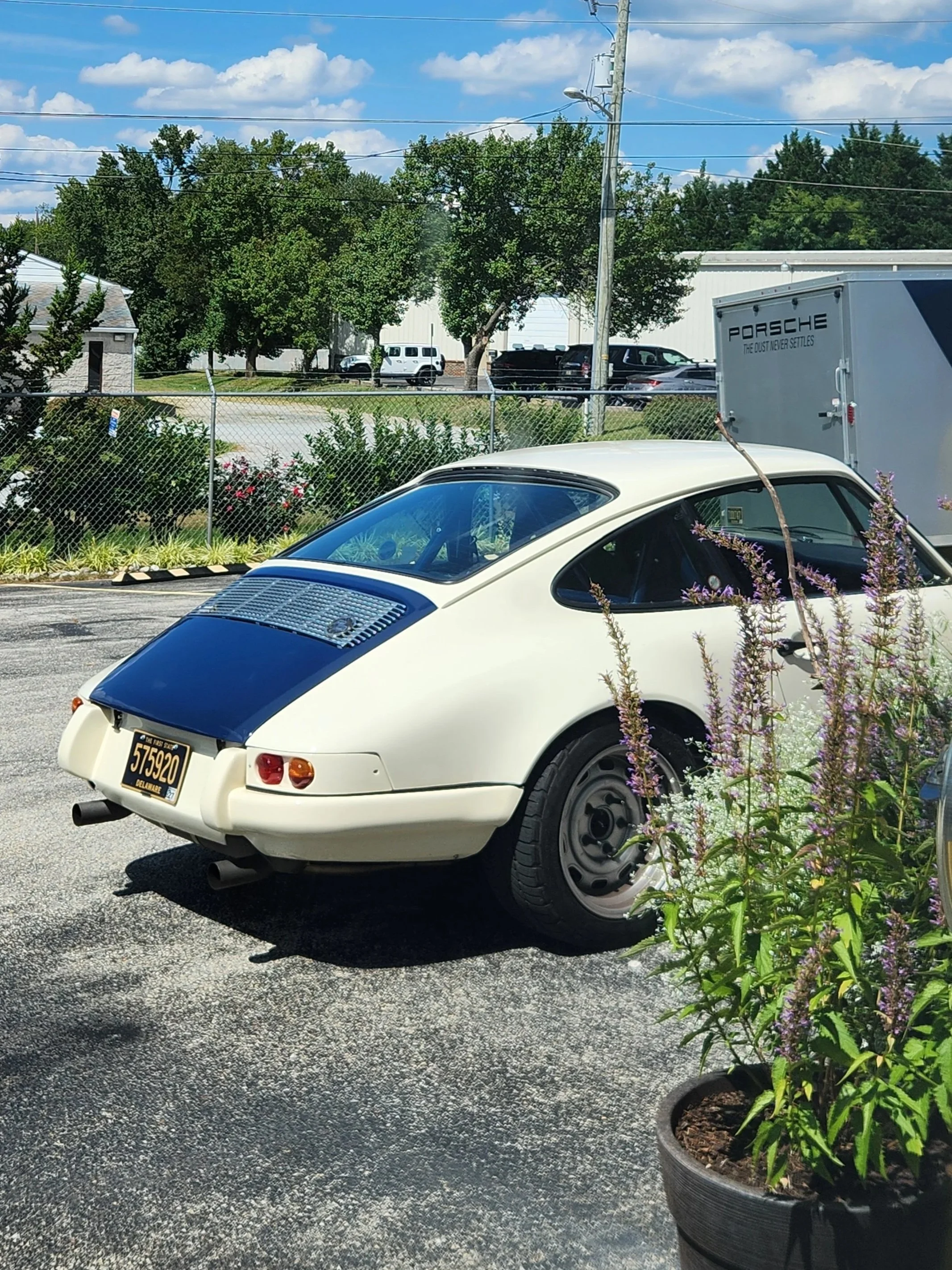 A vintage white Porsche with a blue rear deck lid, parked in a parking lot near some flowering plants and a chain-link fence. The car has an American license plate and a silver rear wheel rim.