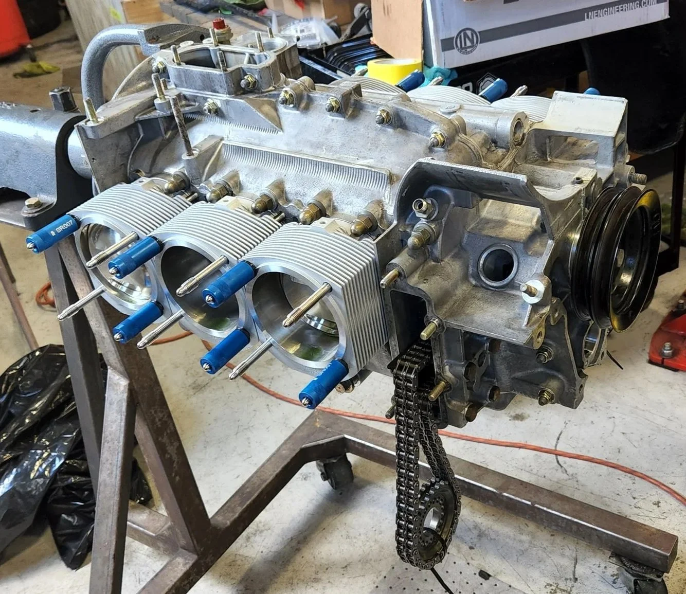 A partially assembled Porsche aircooled engine on a stand, featuring six cylinders with blue hold downs and a timing chain hanging out, in a workshop setting.