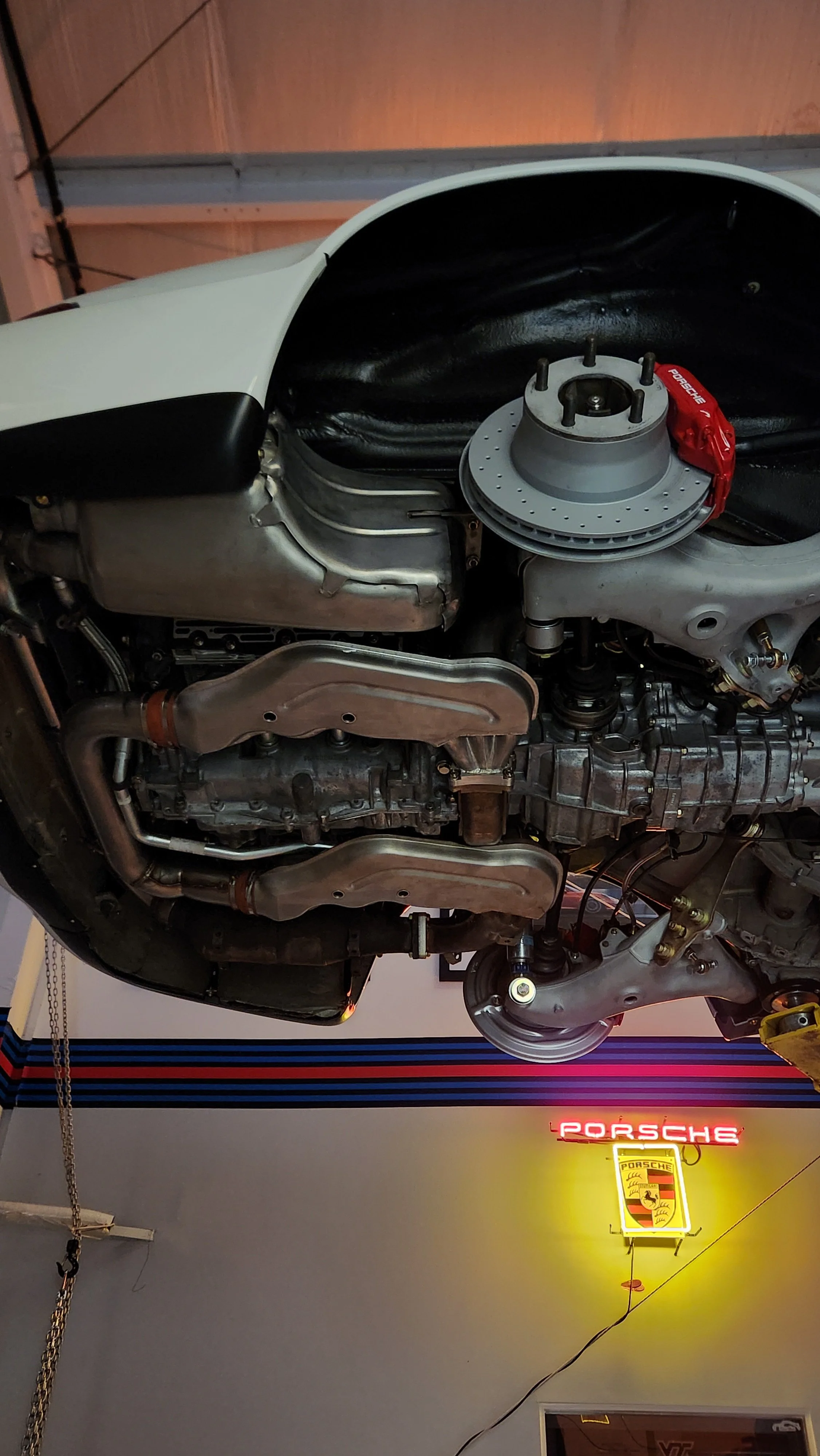 Upside-down view of a classic Porsche 964 engine and mechanical components in a garage with a neon Porsche sign on the wall.