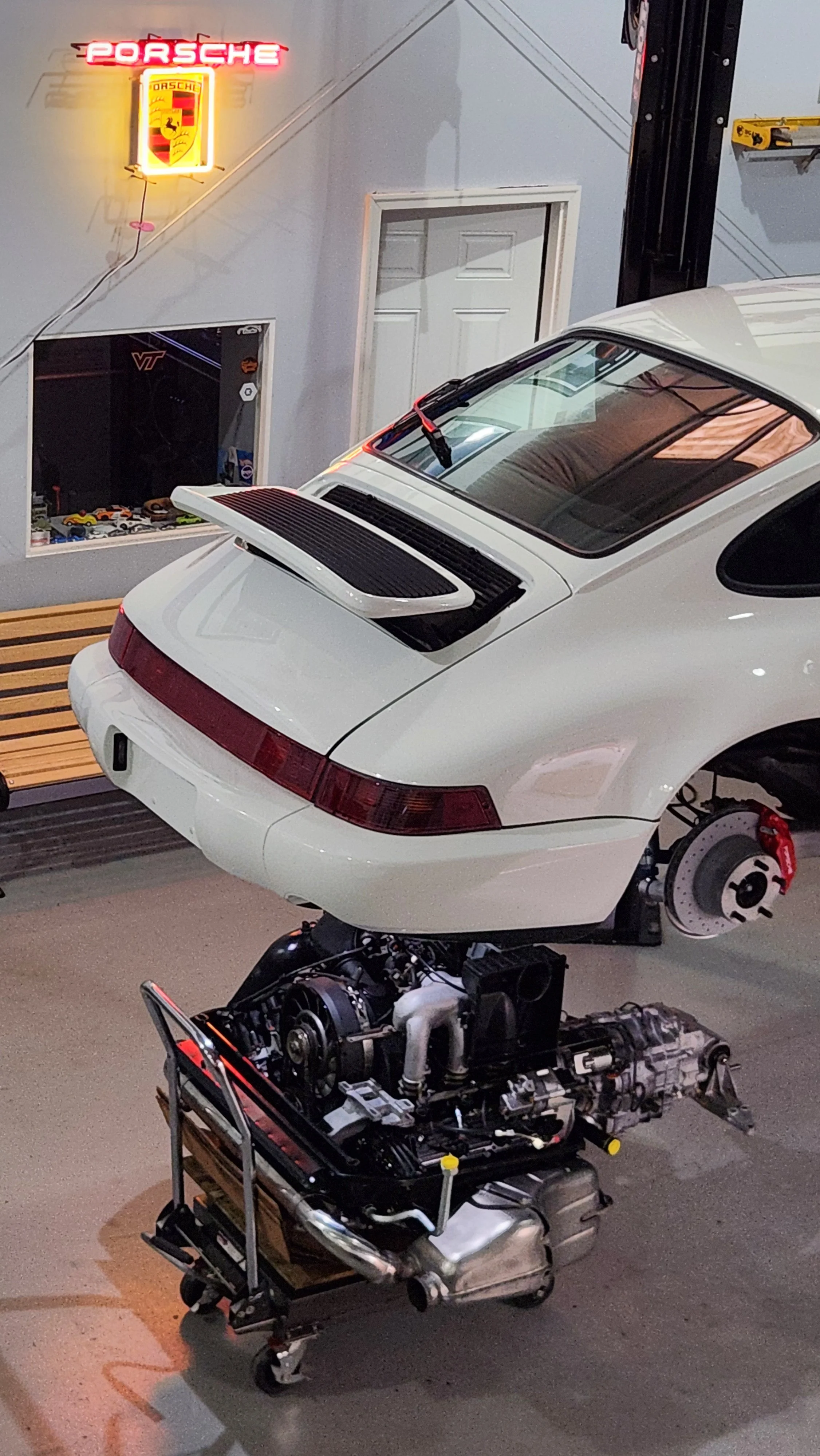 A white Porsche 911 with its engine removed, shown in a garage or workshop. The car is elevated, and the engine is placed on a cart nearby. The garage has Porsche and Virginia Tech decorations.