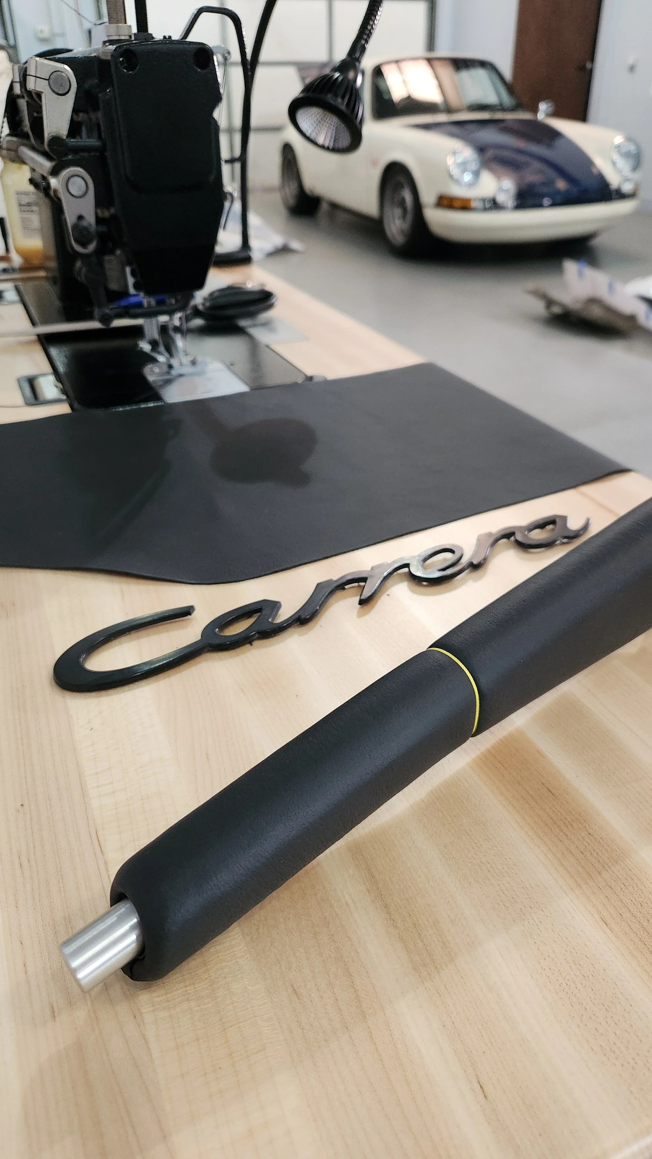 Close-up of a sewing machine on a wooden table with a Porsche Carrara logo and a black leather parking brake handle with yellow stich. A classic white and blue Porsche car is in the background in a workshop or garage setting.