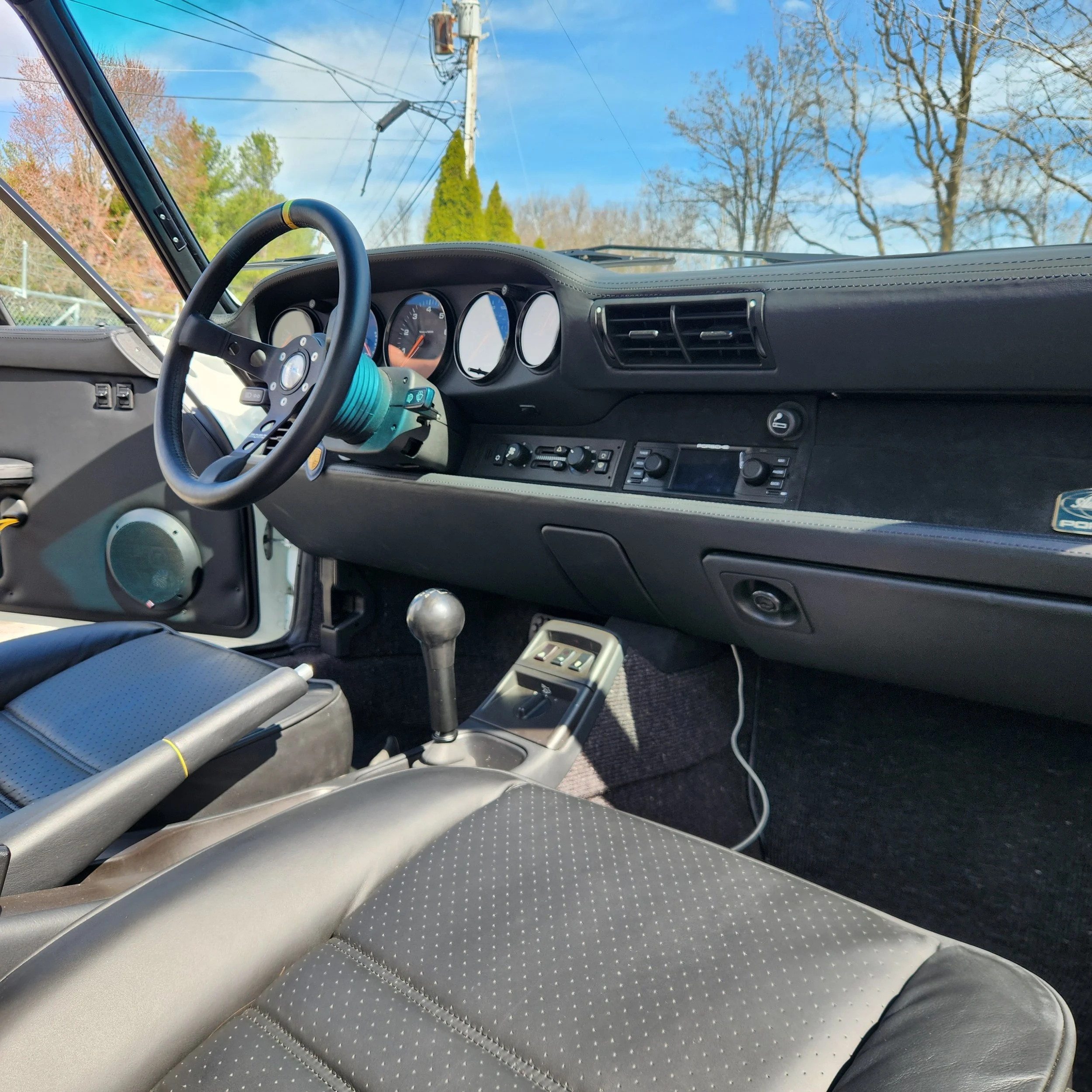 Interior of a vintage car dashboard with steering wheel, gear shift, black seats, and clear view of the road outside with blue sky and trees