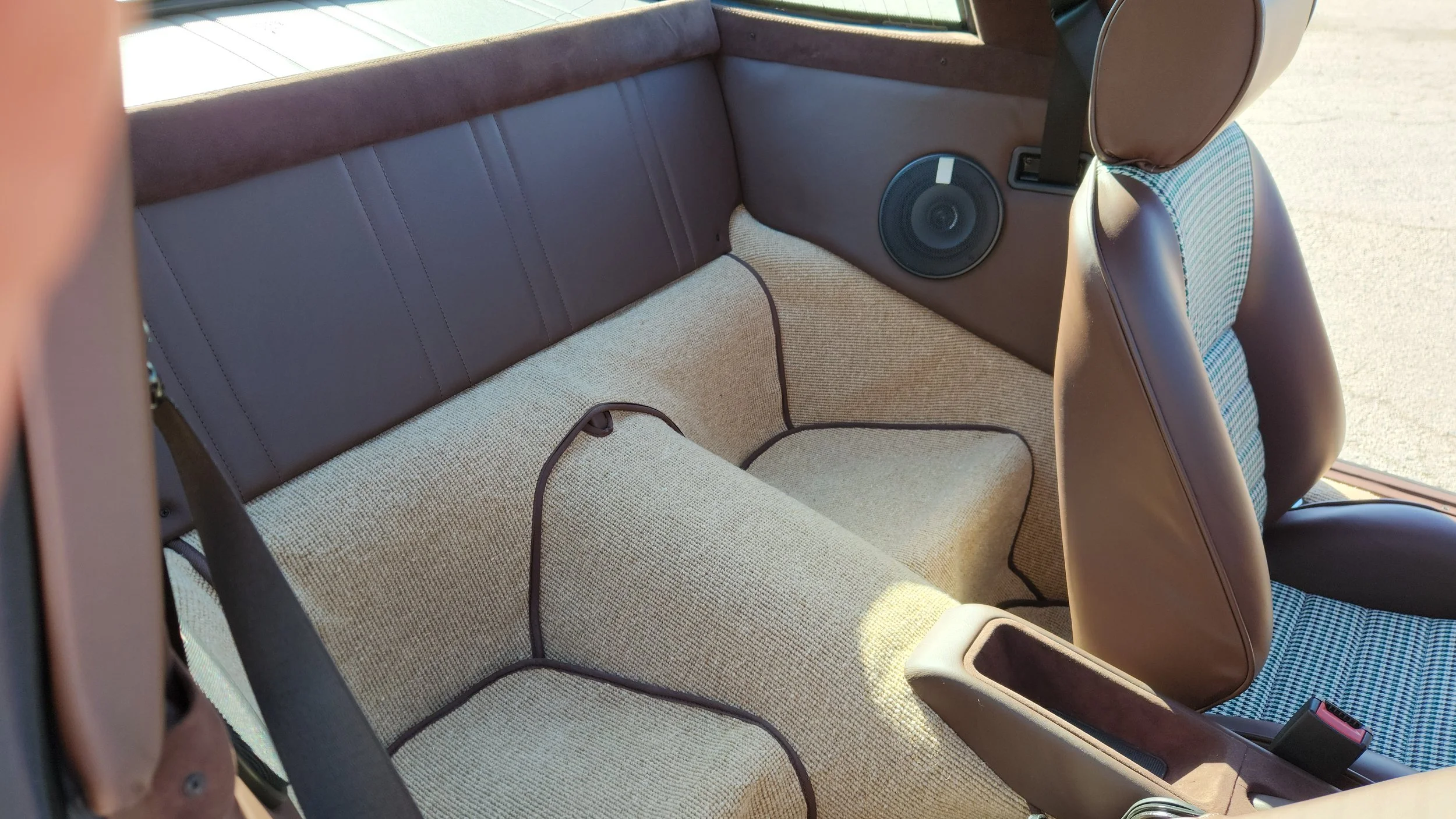 Interior of a vintage 964 Porsche showing the backseat area with brown upholstery, a speaker on the side, and beige carpeted German square weave flooring.
