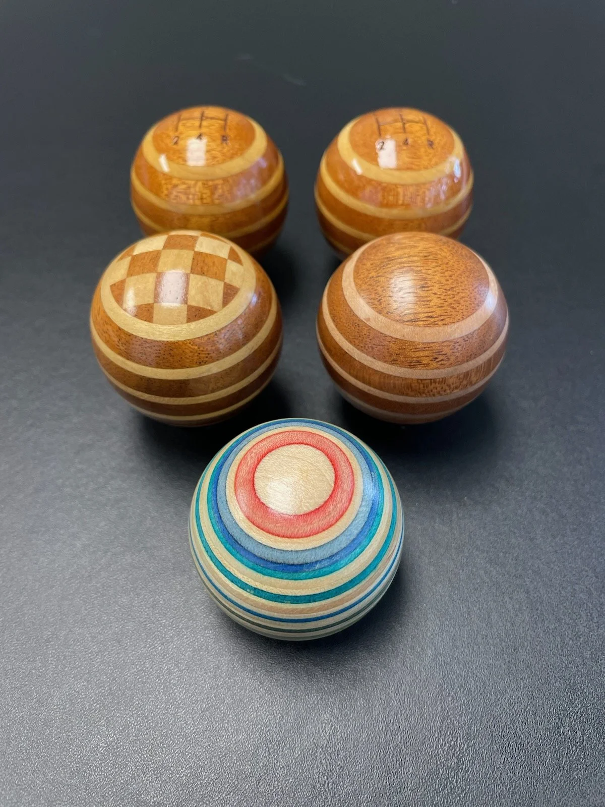 Five wooden shift knobs with various geometric and colorful striped patterns arranged on a dark surface.