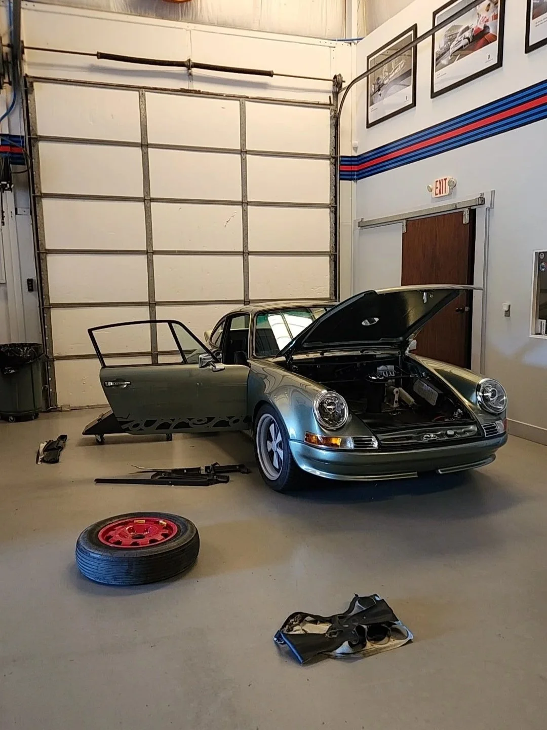 A vintage green Porsche sports car inside a garage with the hood open and the driver side door open. Car parts and tools are scattered on the floor, including a wheel with a red rim, a tire, and some black mechanical parts. The garage has white walls decorated with framed pictures, a large closed garage door, and an exit door with an emergency exit sign above it.