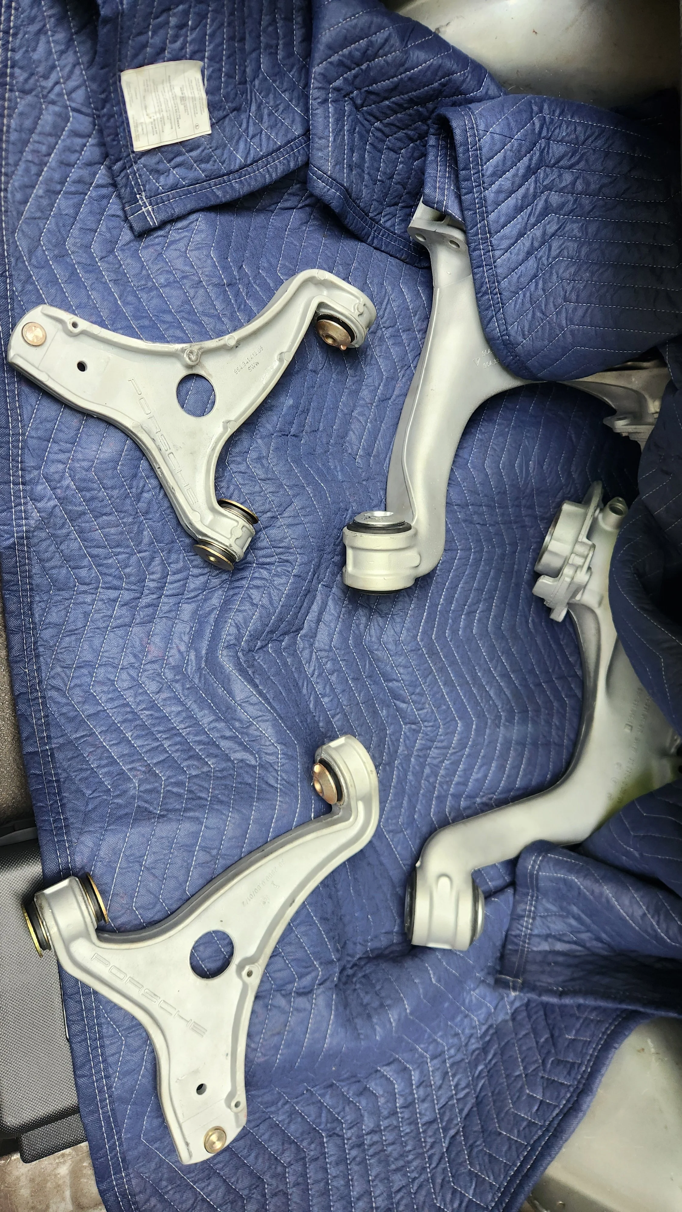 Two silver Porsche control arm and two trailing arm suspension parts with rubber bushings lying on a blue quilted blanket.