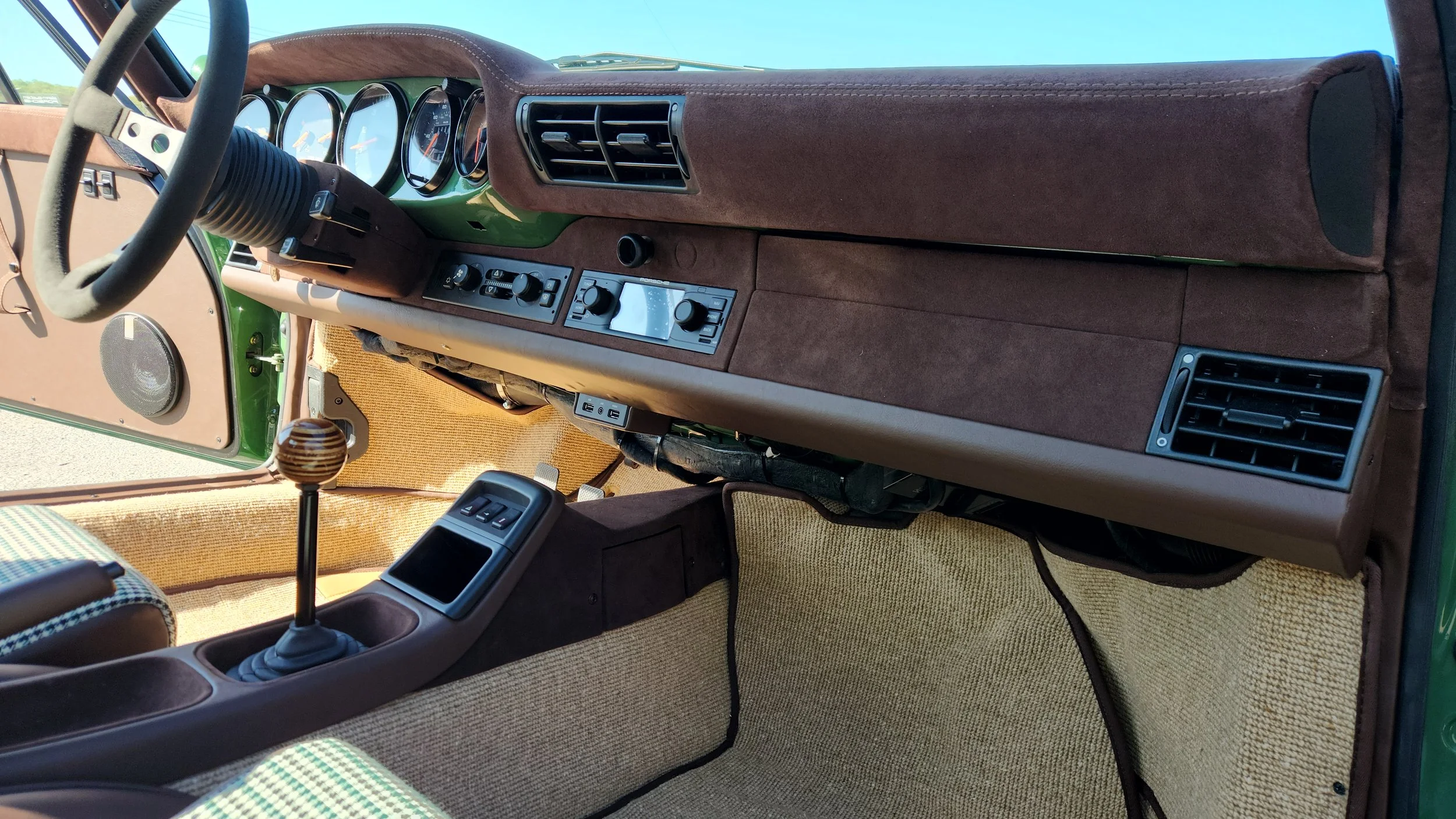 Interior of a vintage car showing a brown and green dashboard with analog gauges, air vents, a gear shift, and a beige and checkered seat.