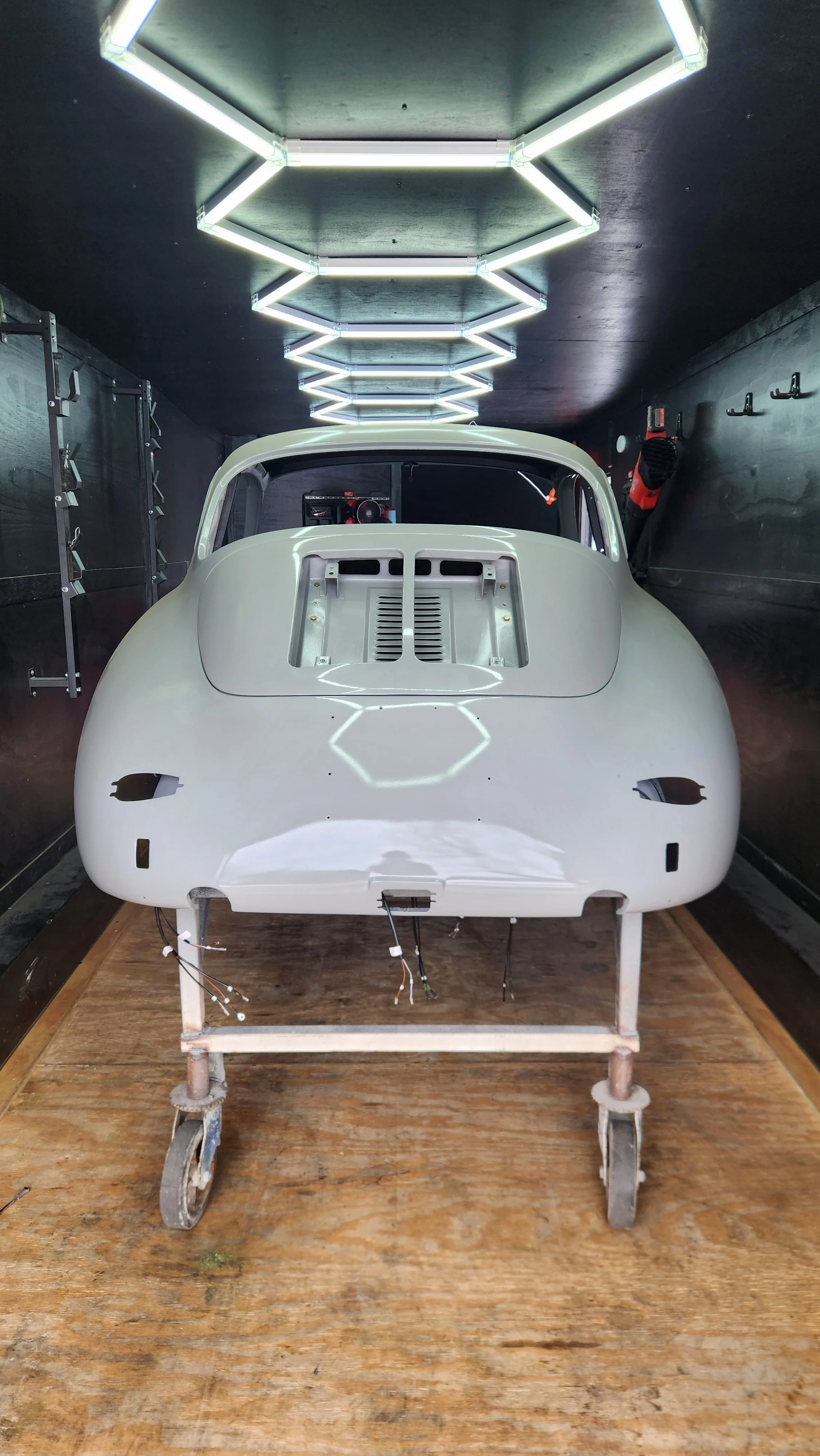 A classic grey sports car 356 body shell on a wheeled cart inside a trailer with black walls and illuminated hexagon ceiling lights.