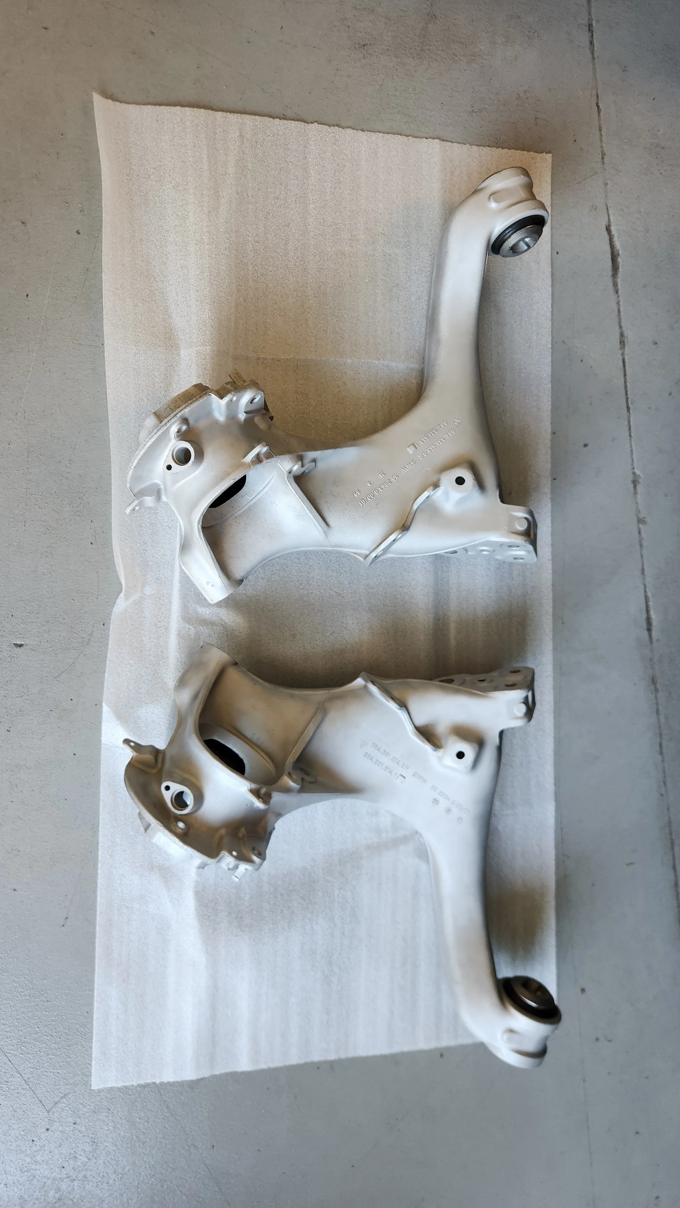 Two silver automotive suspension trailing arms placed on a piece of white foam on a concrete floor.