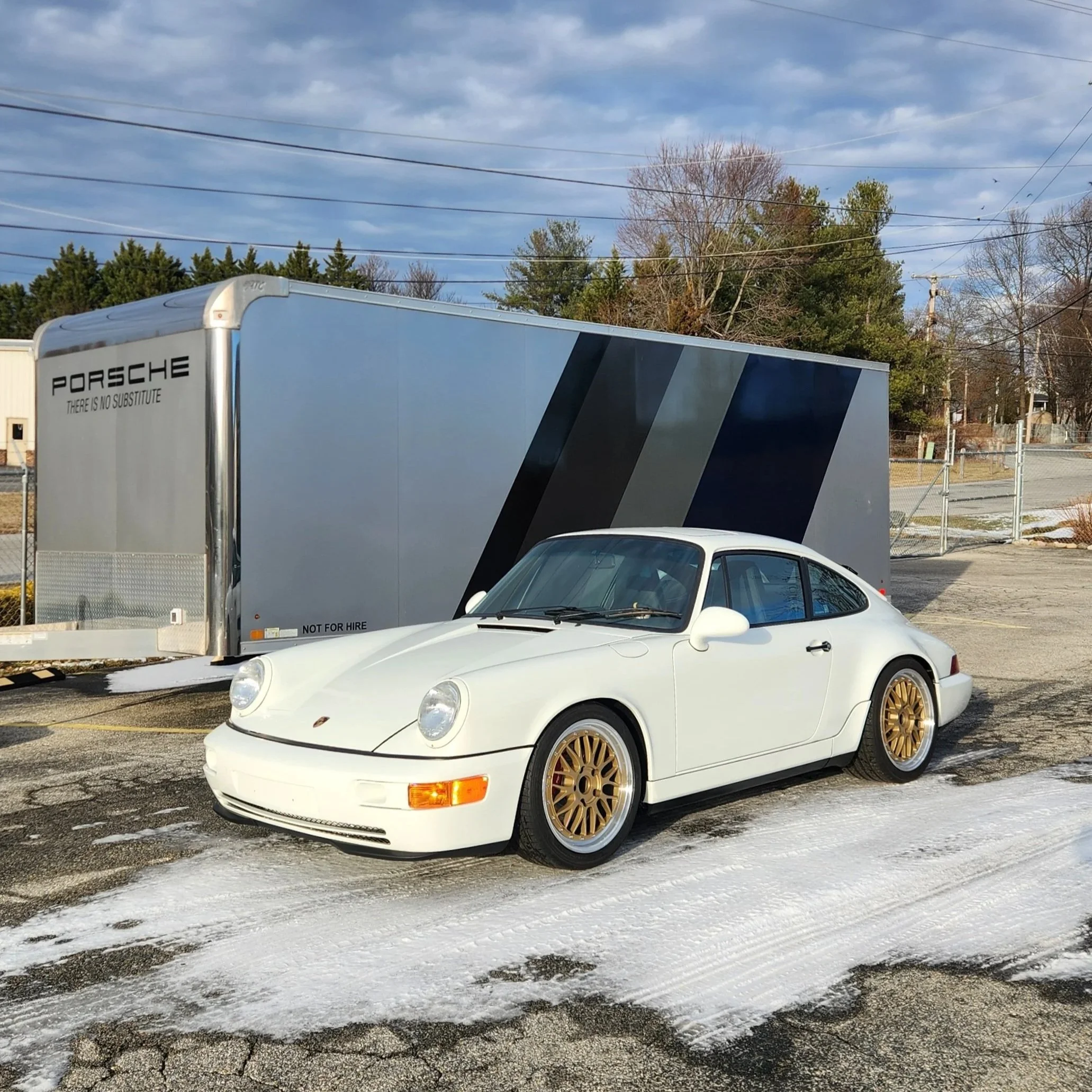 A white Porsche sports car with gold wheels parked on snow-covered ground, with a Porsche trailer in the background and a partly cloudy sky.