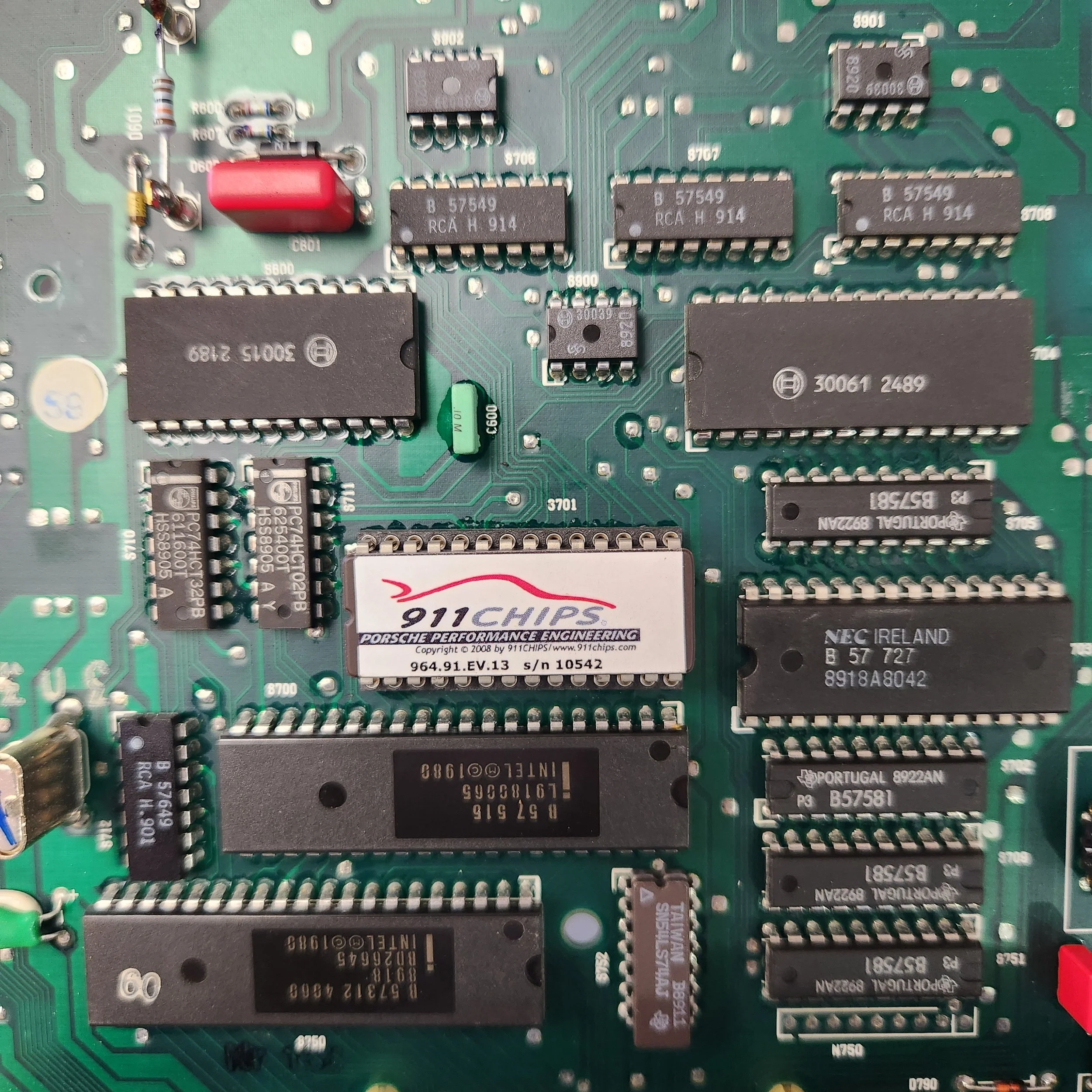 Close-up of a green electronic circuit board with various integrated circuits, resistors, and connectors, including a Porsche 911 Performance Engineering chip in the ECU