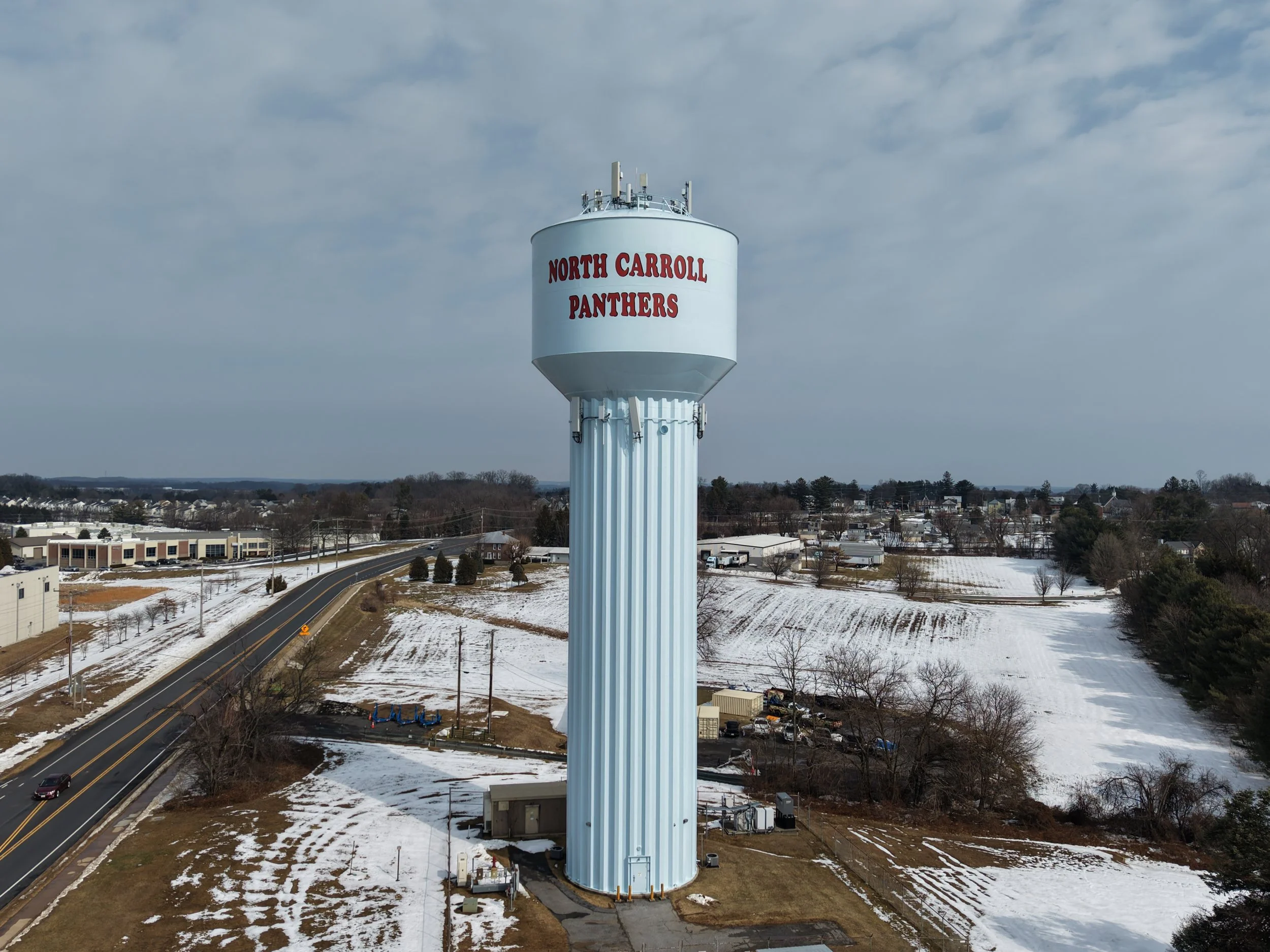 A water tower with the words 'North Carroll Panthers' written on it, located in a semi-rural area with snow-covered fields and a road nearby.