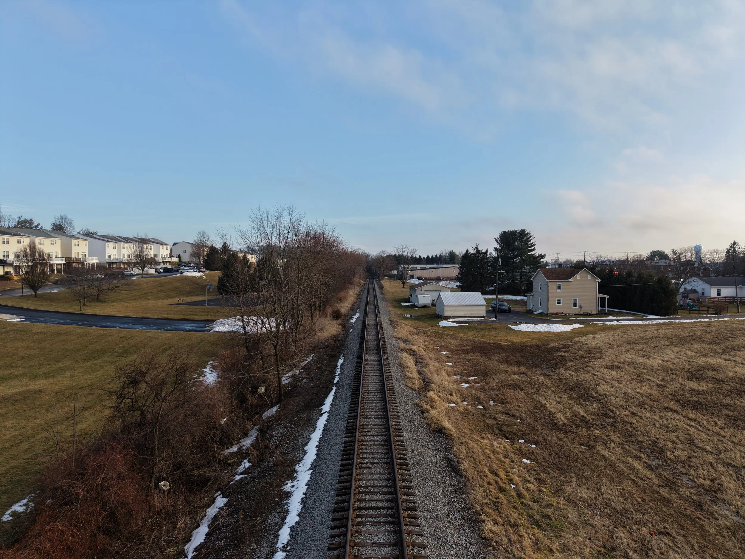 A straight train track runs through a suburban landscape with houses, trees, and patches of snow on either side, under a hill sky.