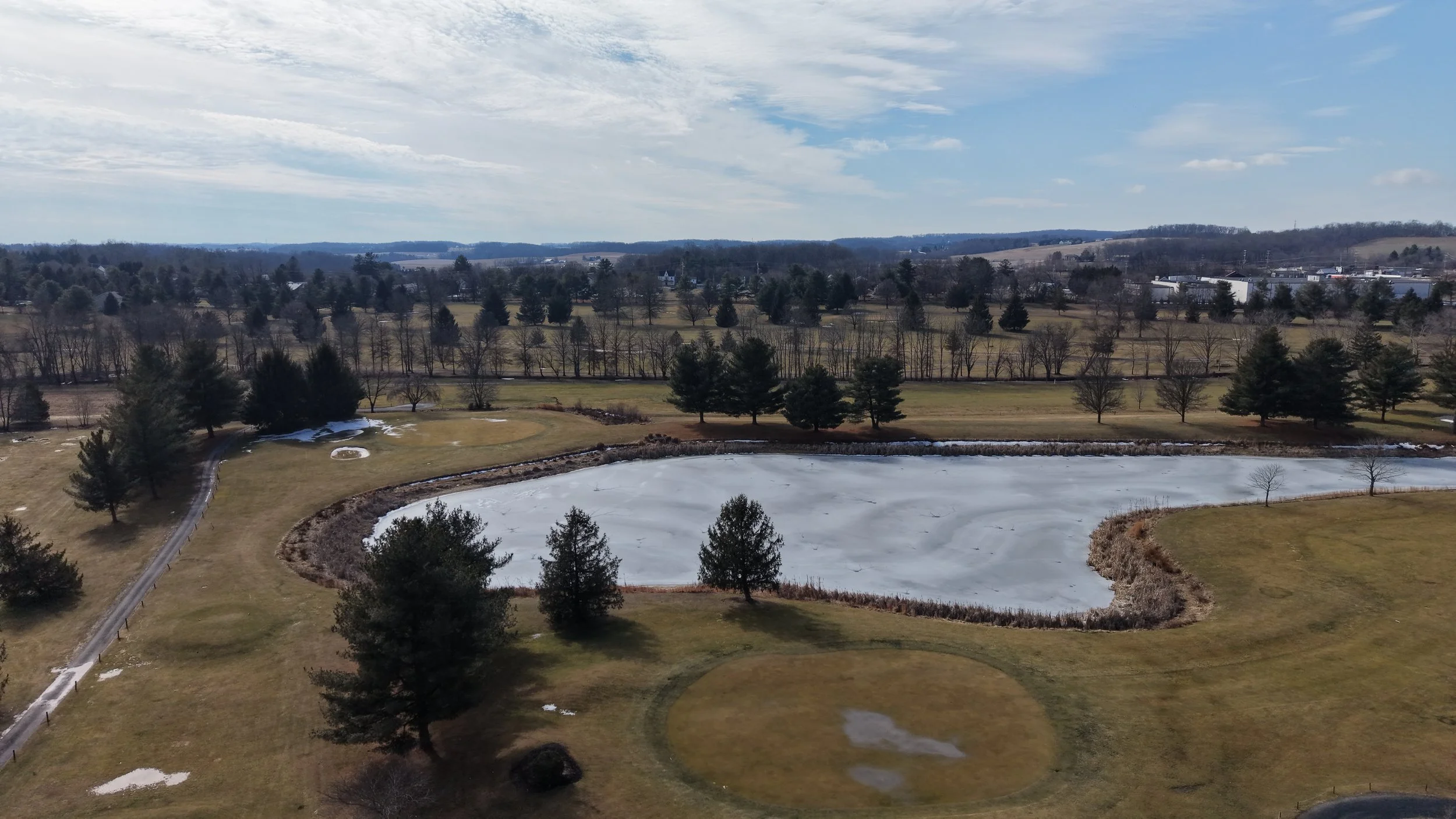  aerial view of a partially frozen pond surrounded by grassy areas and trees in a rural landscape under a partly cloudy sky