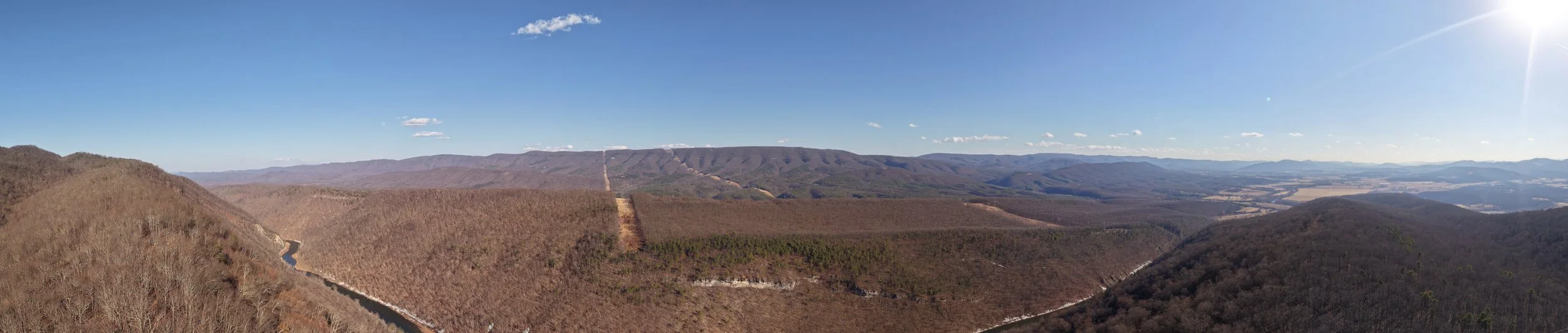 A panoramic view of mountain ranges covered in leafless trees, with a clear blue sky and a bright sun shining from the upper right corner.