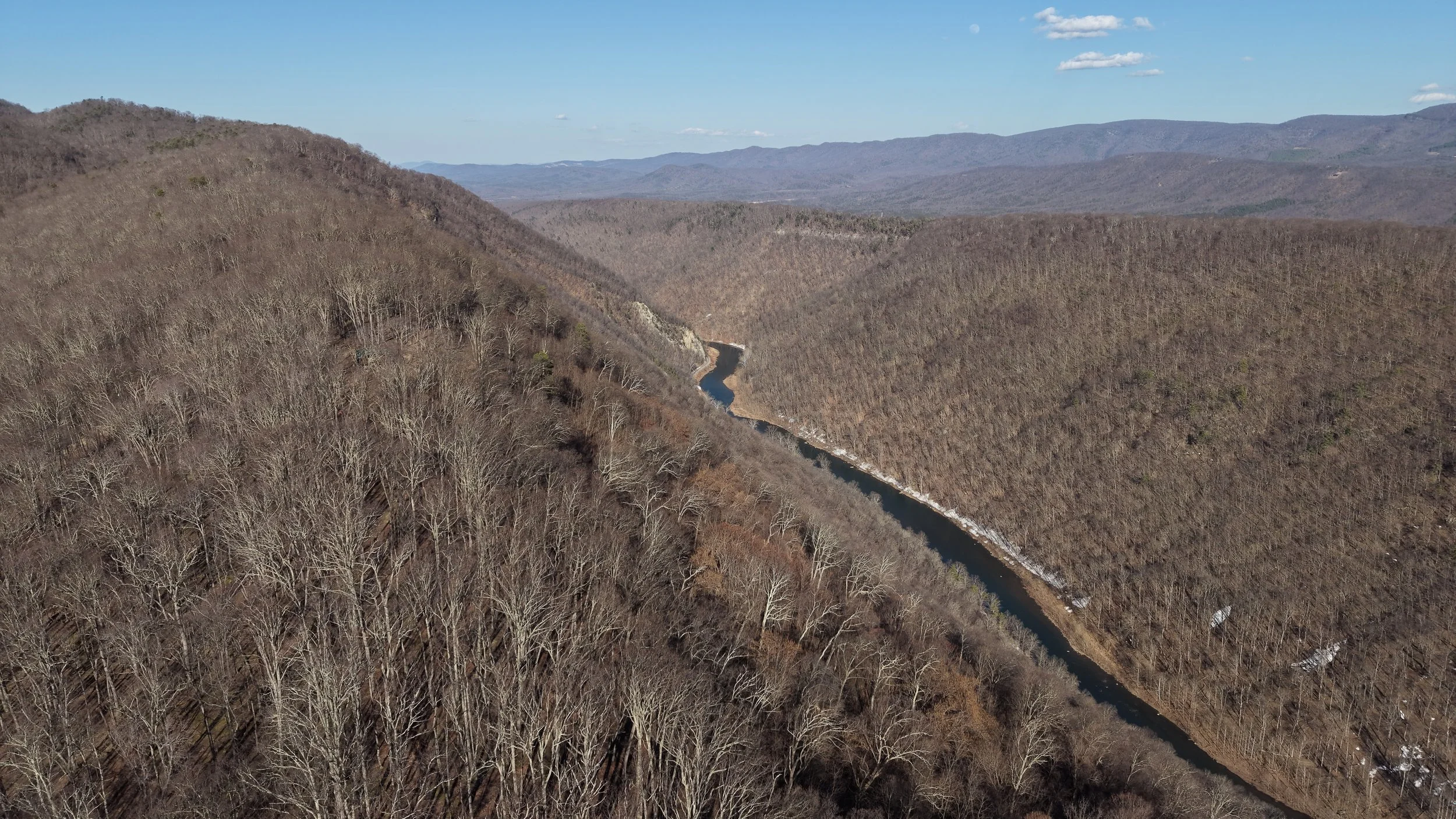 Aerial view of a winding river flowing through a valley surrounded by barren trees on hills and mountains in the distance under a partly cloudy sky.