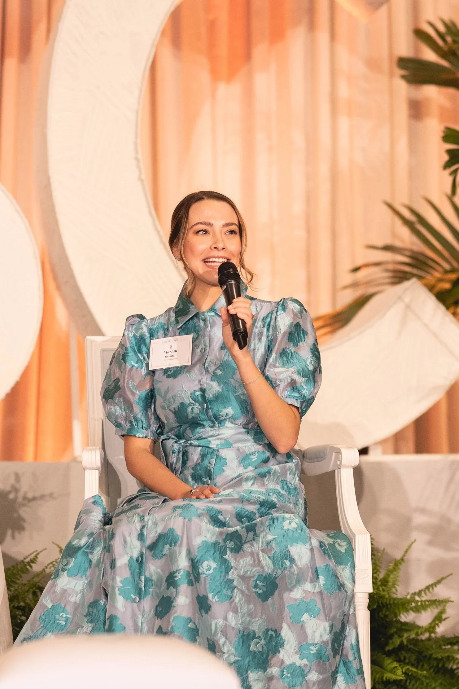 A woman seated on a white chair, holding a microphone, speaking at an event with a nature-inspired backdrop and lush greenery.
