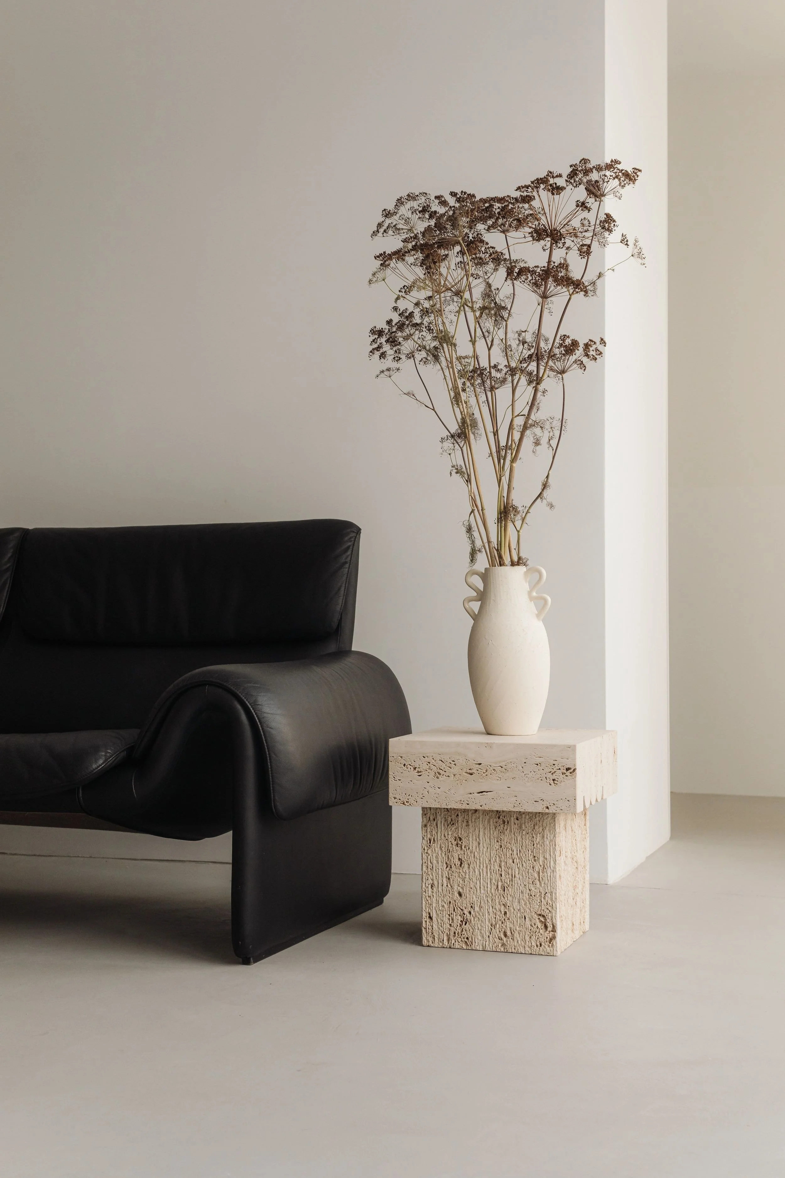 A black leather sofa next to a beige textured pedestal table with a white ceramic vase holding dried branches, set against a minimal white interior.