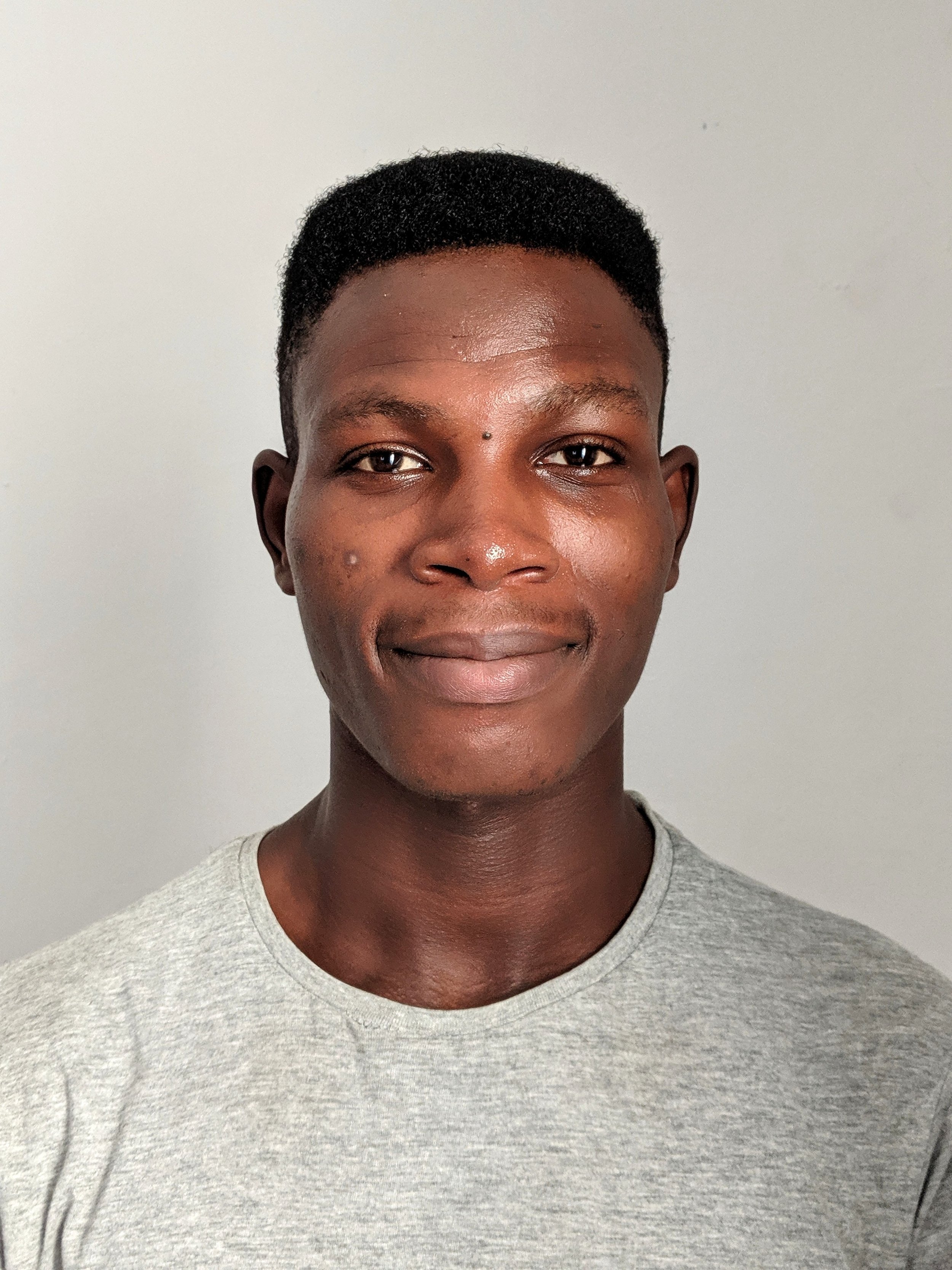 A young man with dark skin, short curly hair, and a slight smile, wearing a light gray t-shirt, standing against a plain white wall.