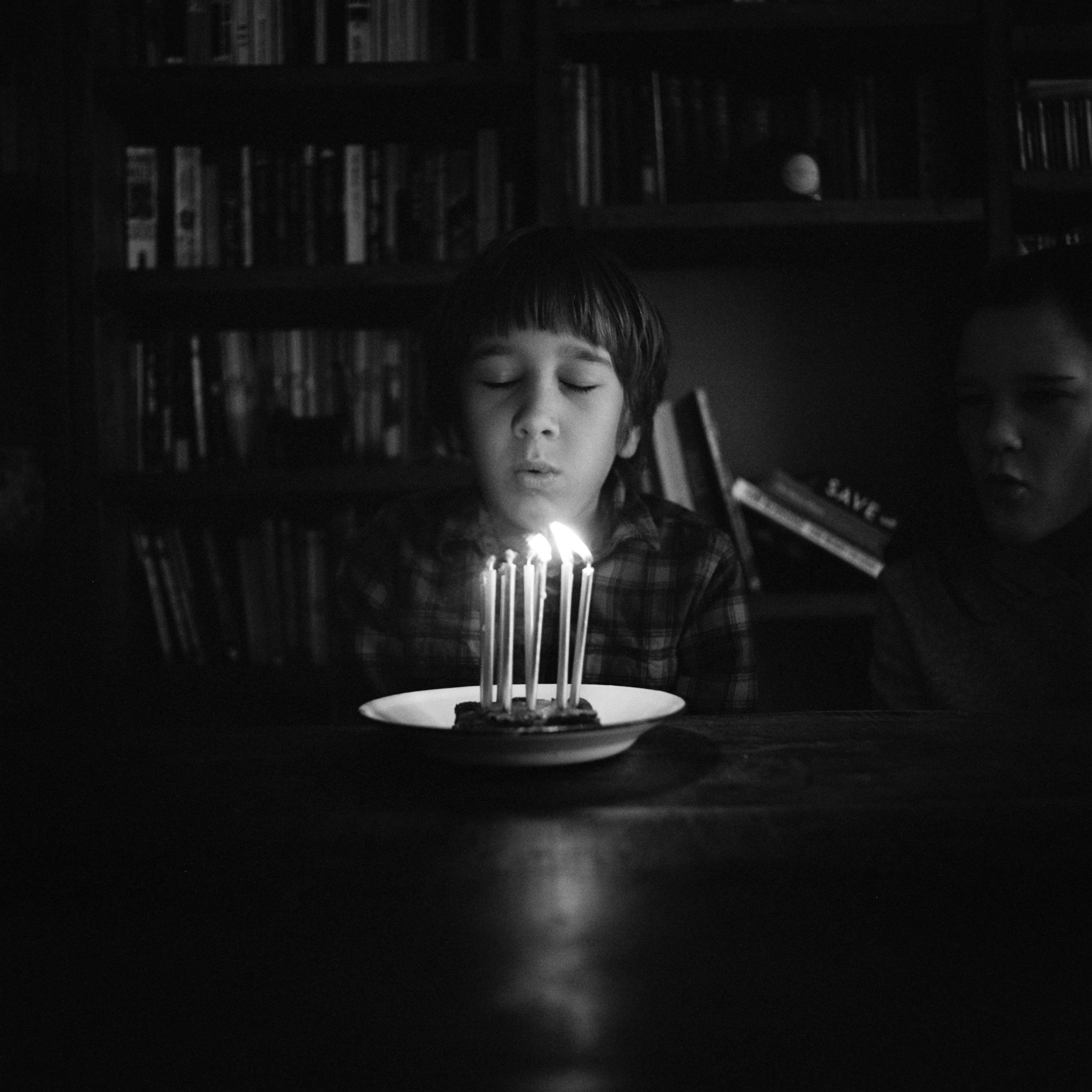 A young boy blowing out birthday candles on a cake in a dark room, with a girl observing nearby.