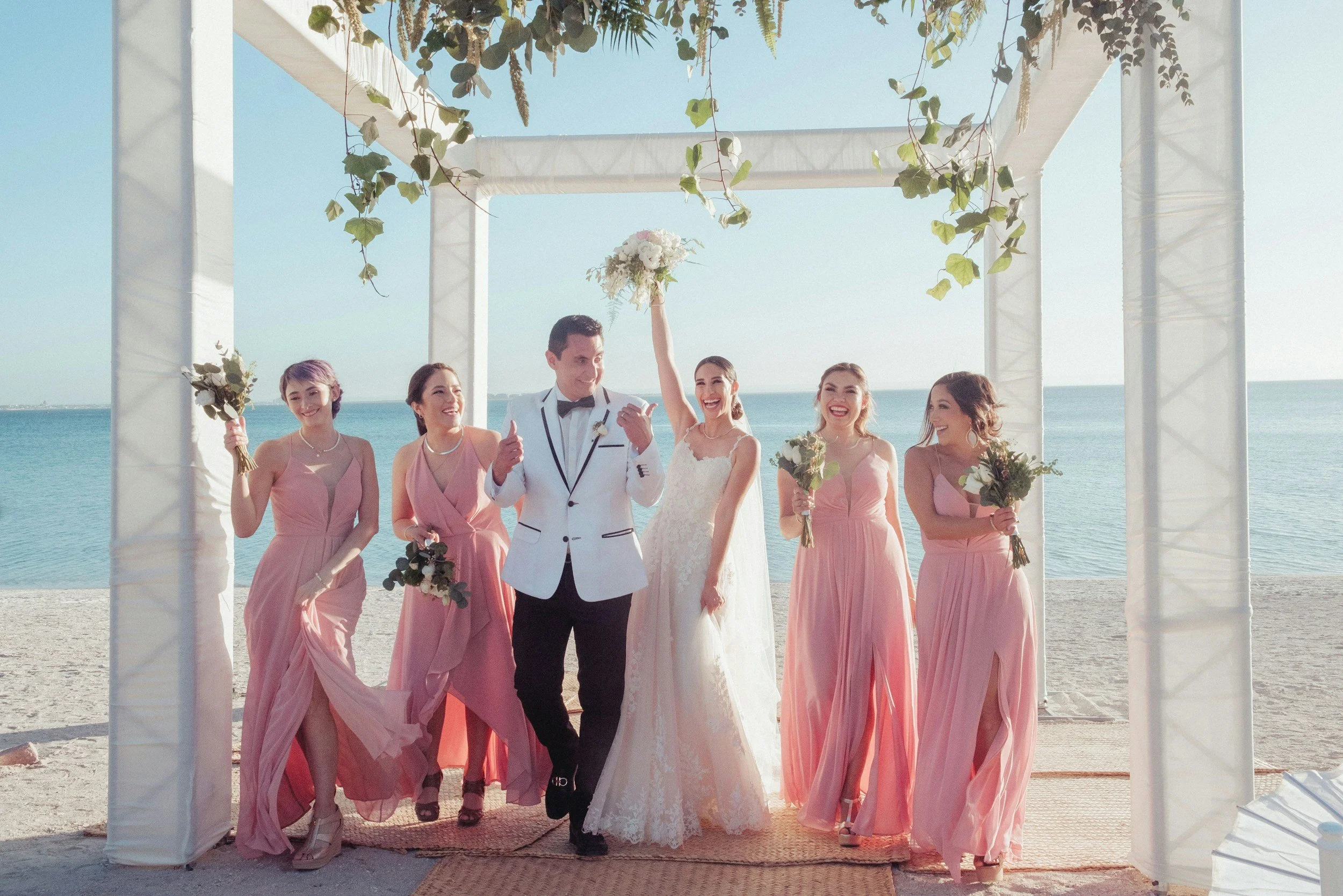 A bride and groom celebrating their wedding on a beach with their bridesmaids. The bride is holding a bouquet and raising her hand, smiling, while the groom stands next to her giving a thumbs up. The bridesmaids are dressed in pink dresses, holding bouquets, and smiling.