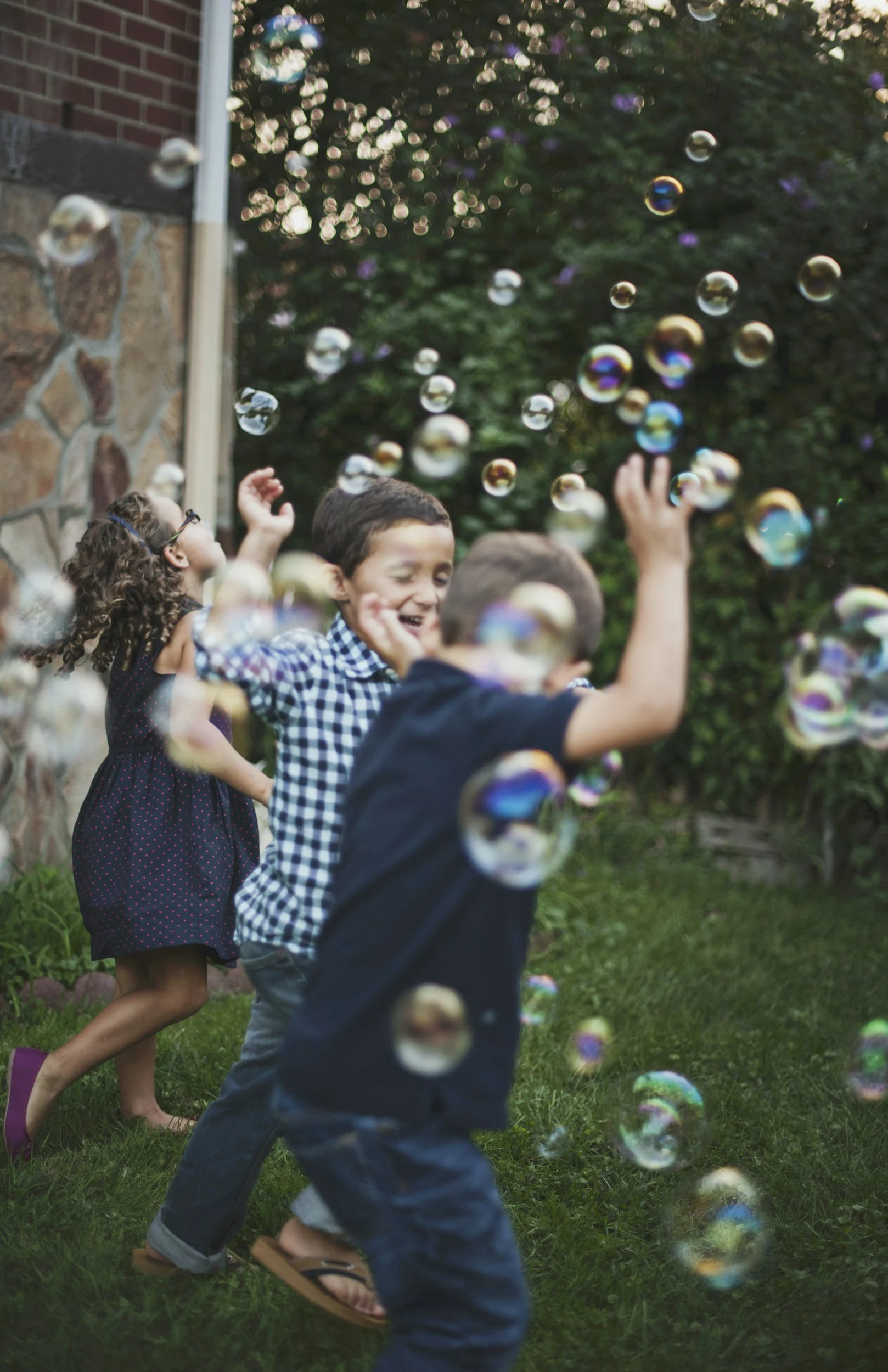 Children playing with soap bubbles outdoors, smiling and reaching for bubbles in a backyard.
