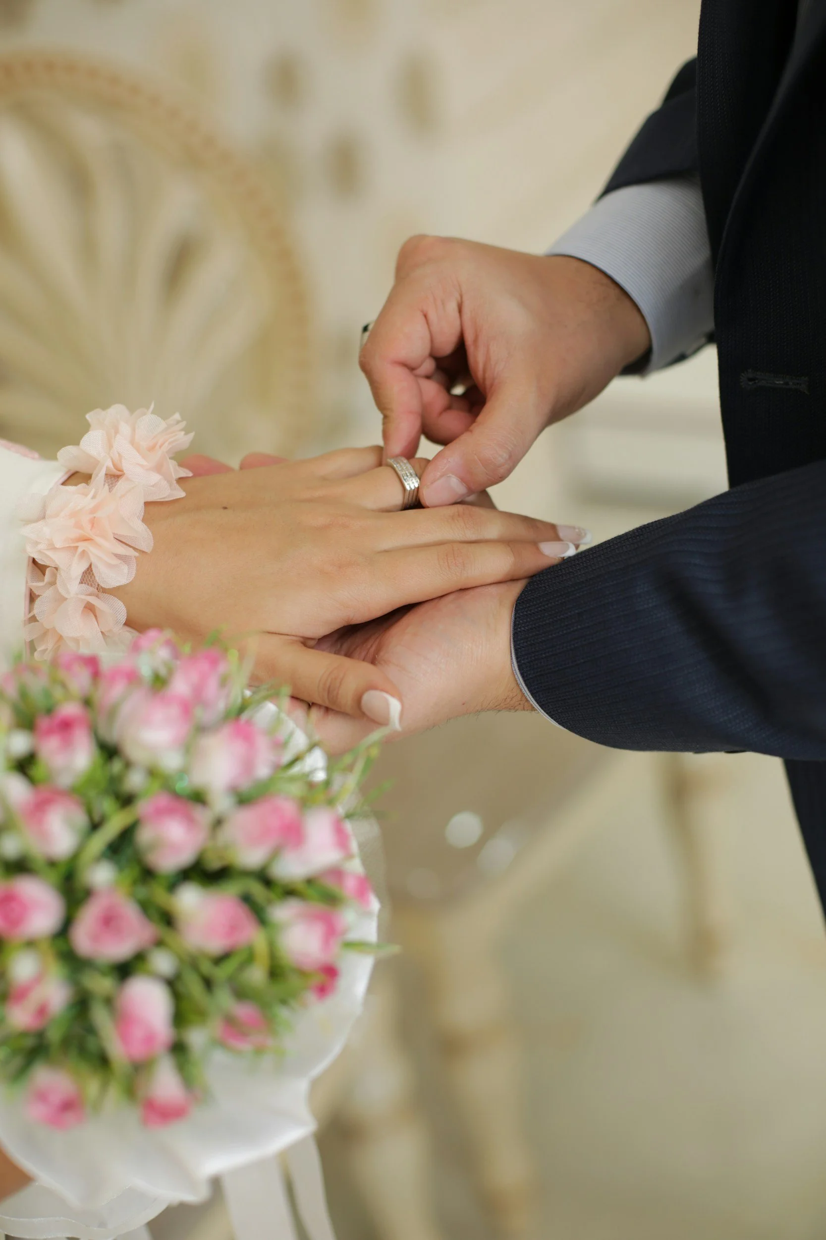 A person is placing a wedding ring on a woman's finger during a wedding ceremony. There is a bouquet of pink flowers in the foreground.
