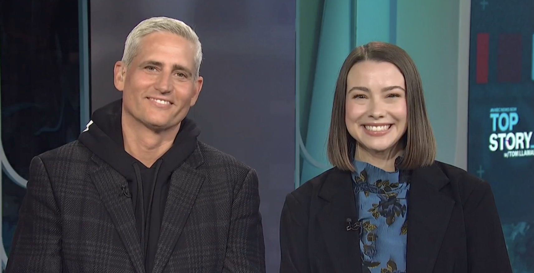 Two news anchors smiling during a TV broadcast, with a studio background including a screen that says 'Top Story'.