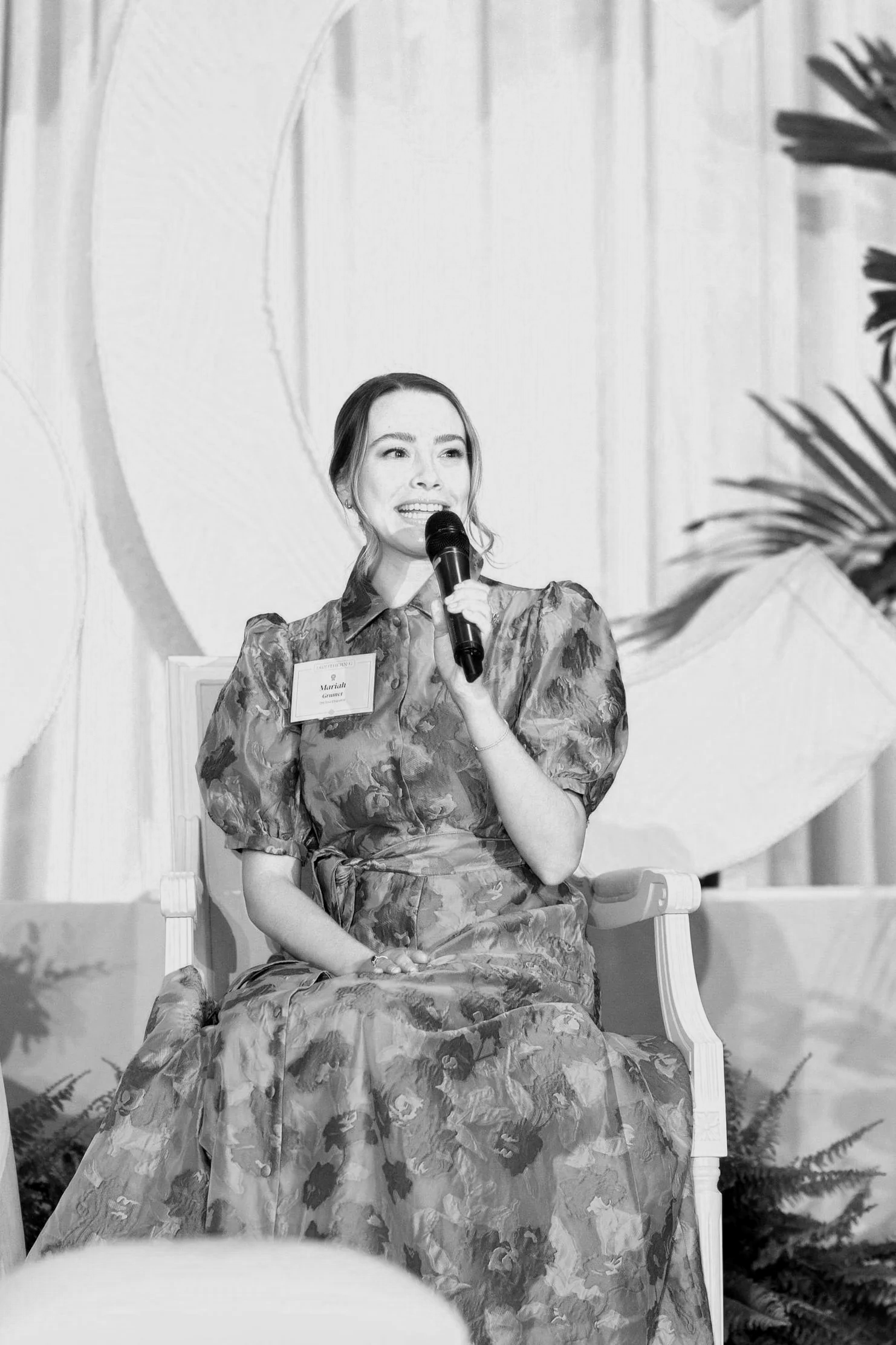 A woman is giving a presentation on stage in a large, modern venue with exposed brick walls. The audience is seated at tables, attentively listening. Two large screens display the same text about imposter syndrome.