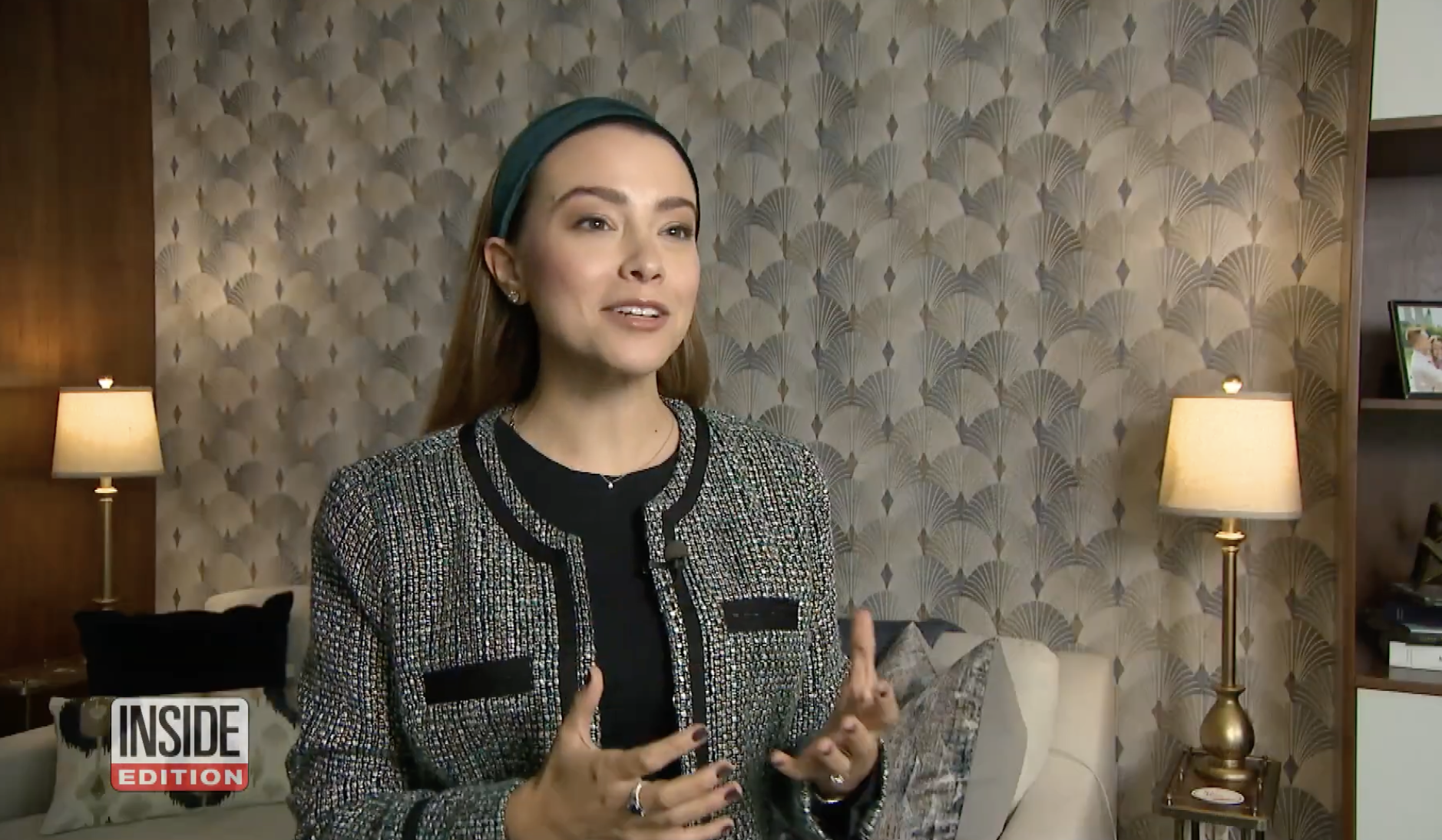 A woman with brown hair, wearing a headband and a sparkly black jacket, is speaking and gesturing with her hands in a living room with patterned wallpaper and two table lamps.