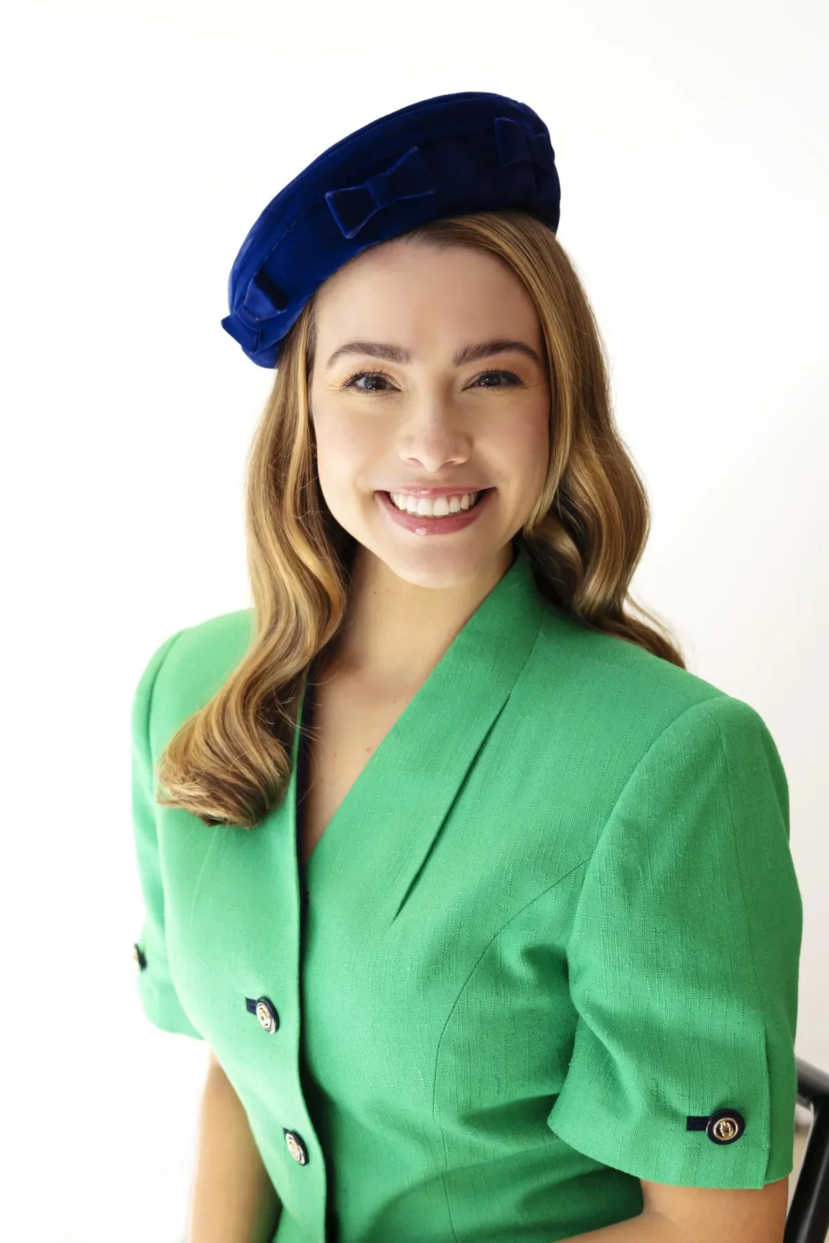 A young woman with long wavy hair smiling, wearing a green blazer and a blue beret with bow detail, standing against a white background.
