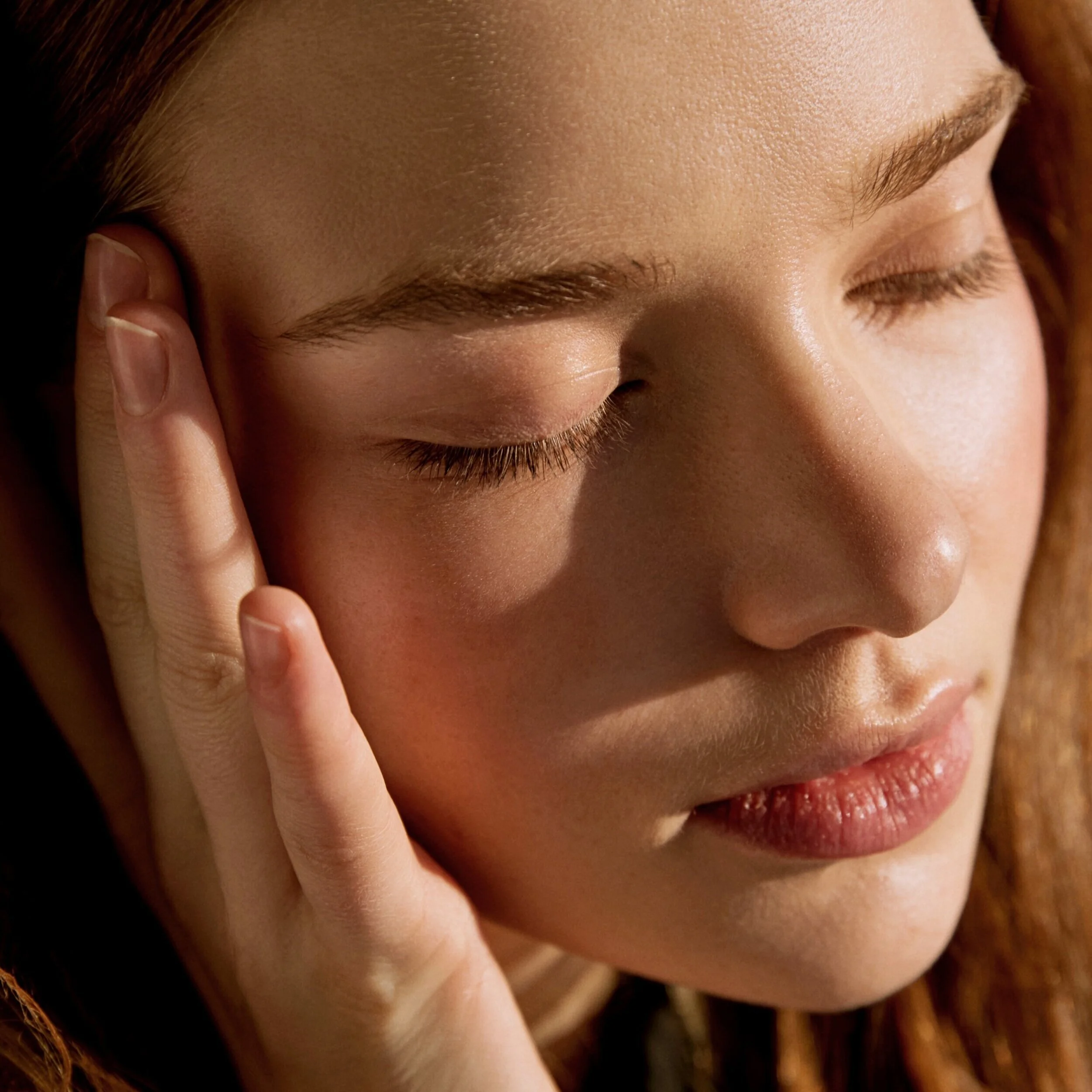 Close-up of a woman with closed eyes, touching her temple with fingers, and relaxed facial expression.