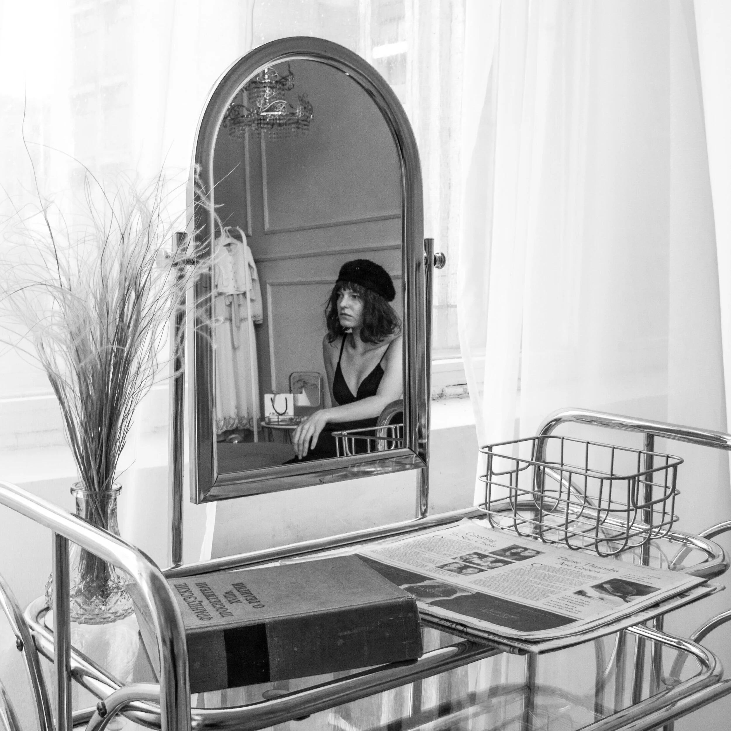 A black-and-white photo of a shopping cart with a large mirror attached to its side, reflecting a woman with brunette hair, wearing a black top and a black beret, sitting in a room with a chandelier, curtains, and wall molding.