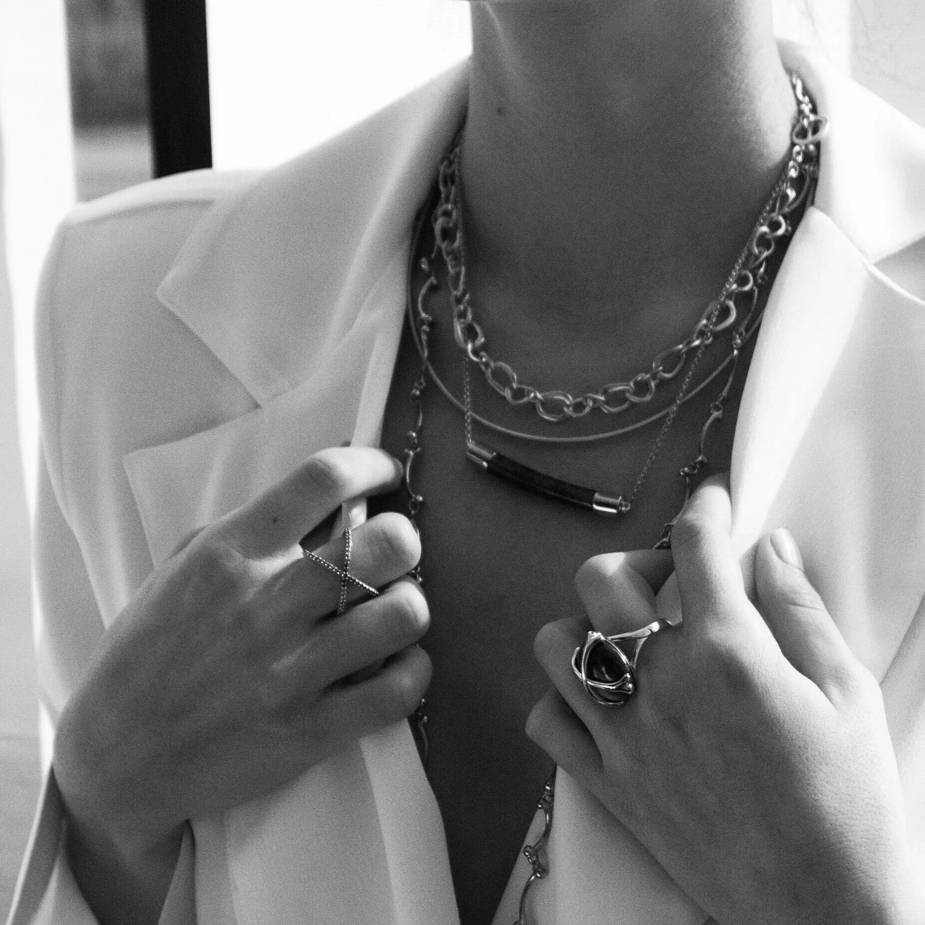 Close-up of a person's neck and chest area, wearing multiple necklaces and rings, with part of a white blazer visible.