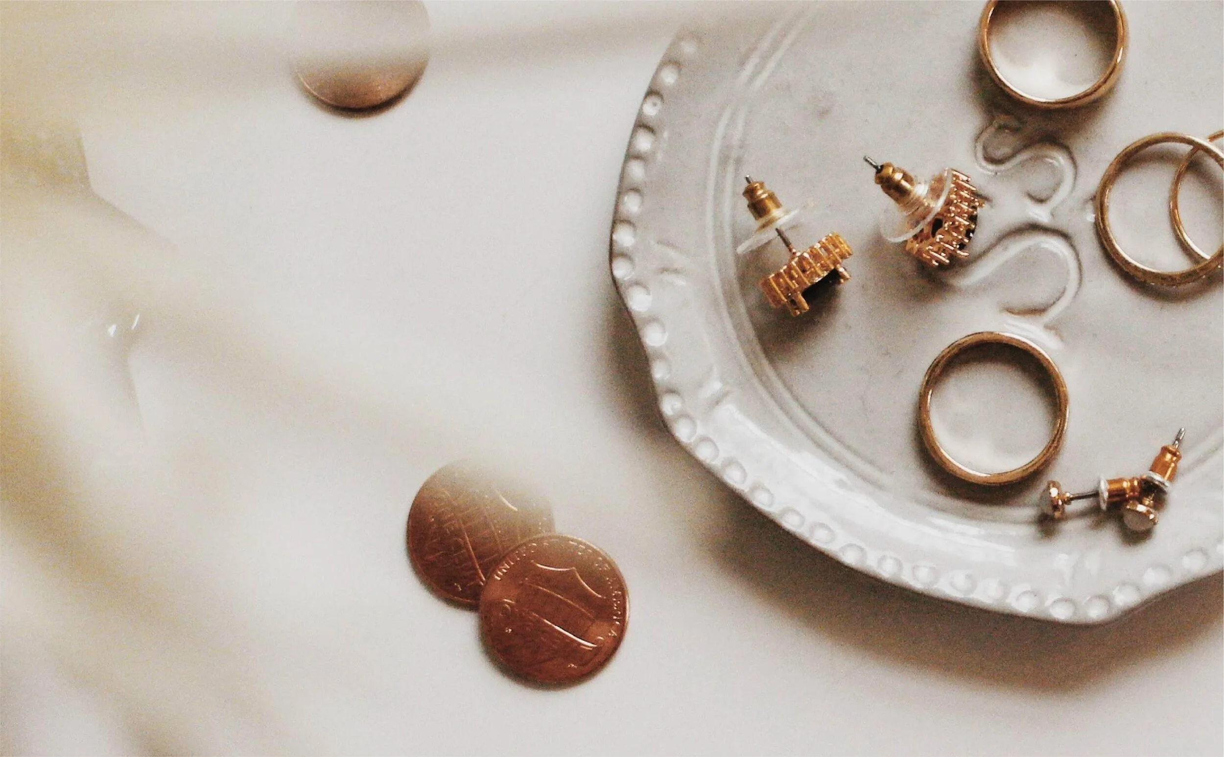 A silver dish with raised decorative edges, holding gold and clear rhinestone earrings, surrounded by scattered copper pennies on a white surface.