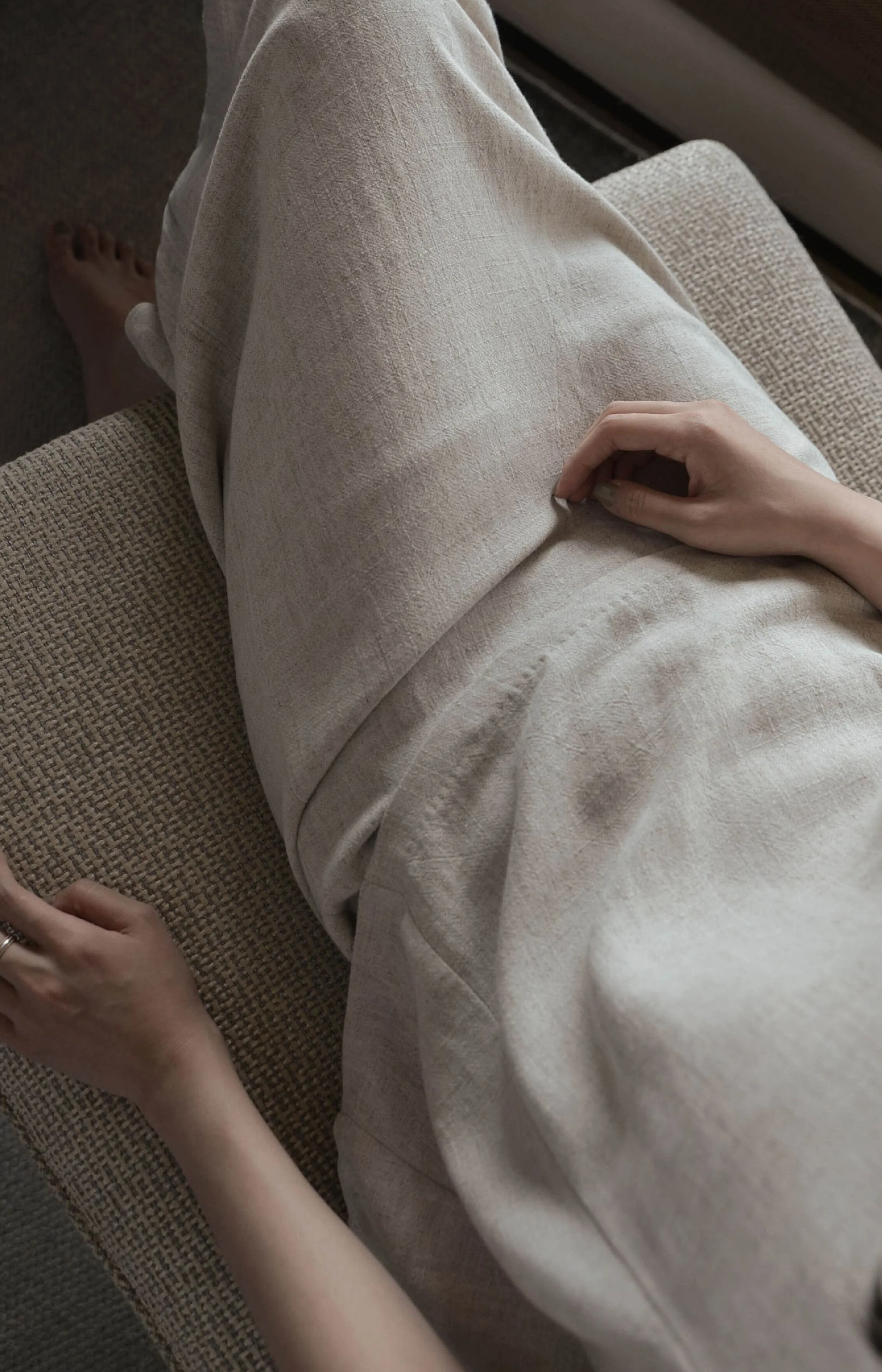 A person sitting on a beige textured couch with their legs crossed, wearing light-colored loose pants, with their left hand resting on their lap and holding a mobile phone, and their right hand on their knee.