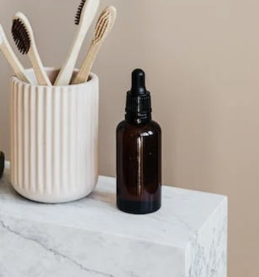 A white ribbed ceramic cup holding bamboo toothbrushes next to a brown glass dropper bottle on a white marble surface.